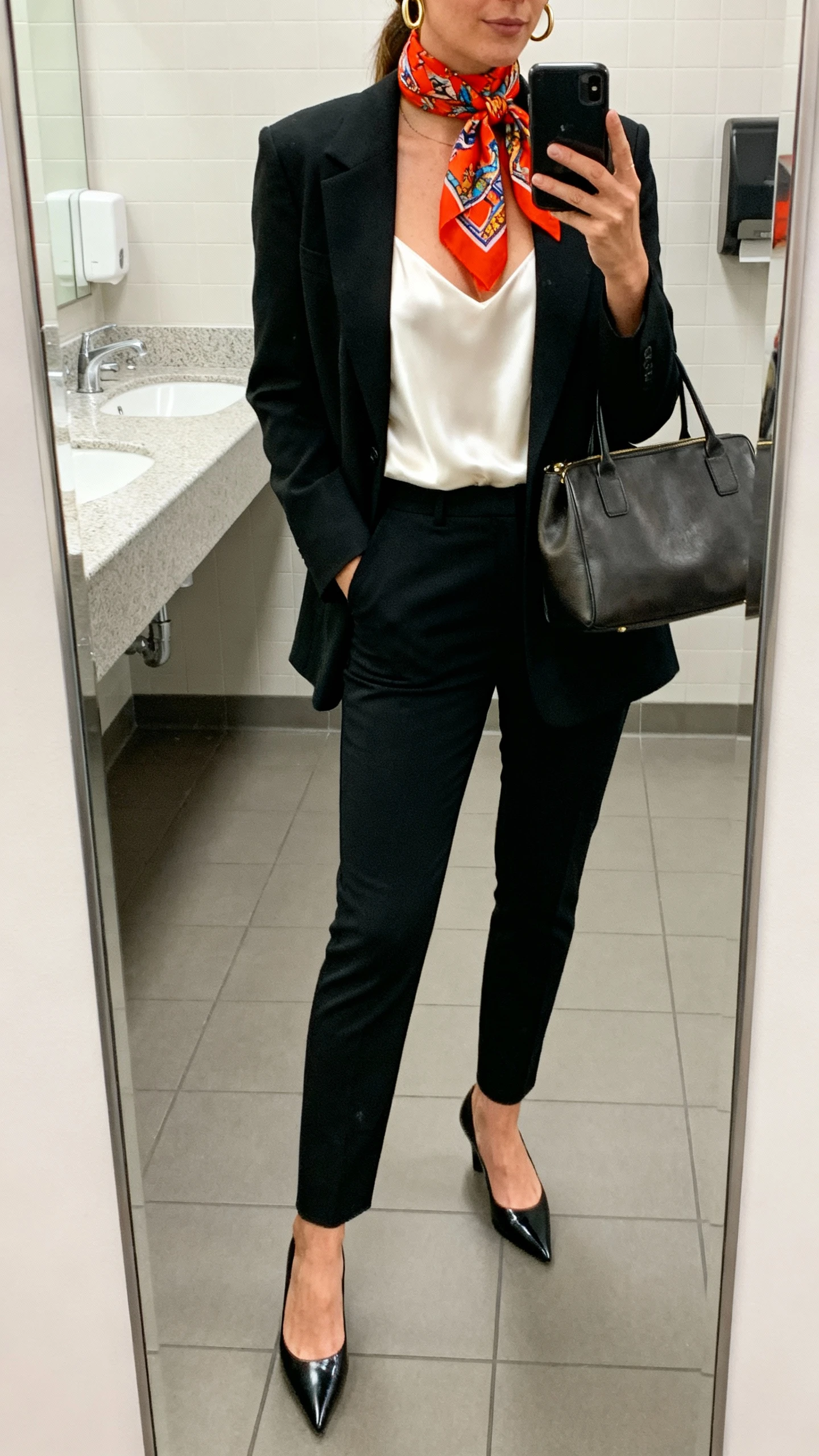 iPhone mirror selfie of a woman wearing a tailored black blazer over a white silk camisole, slim black trousers, pointed-toe heels, a bright patterned scarf knotted at the throat, structured tote bag, and minimalist gold hoops, face not visible, cropped at shoulders, office bathroom mirror, indoor lighting, iPhone photo quality.