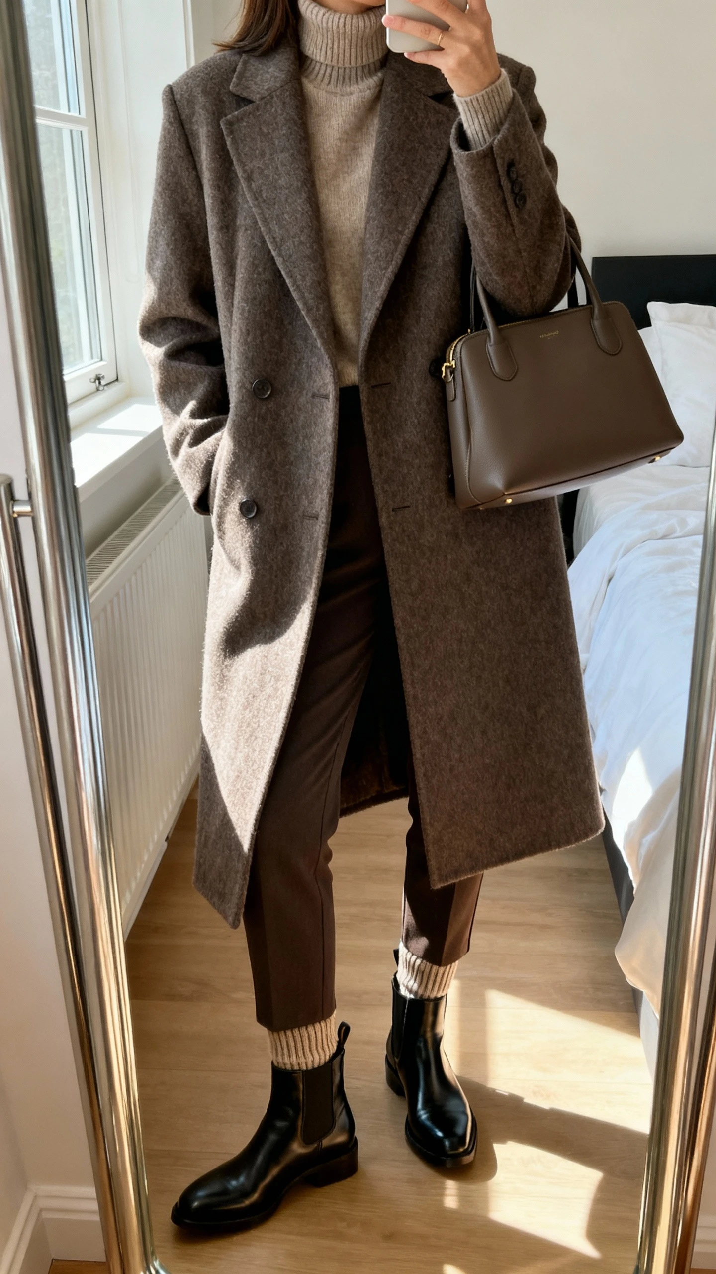 iPhone mirror selfie of a woman wearing a tailored wool coat over a cashmere turtleneck, slim ankle trousers, sleek Chelsea boots with luxe ribbed socks, structured tote, face not visible, cropped at shoulders, bedroom mirror, soft window light, iPhone photo quality.