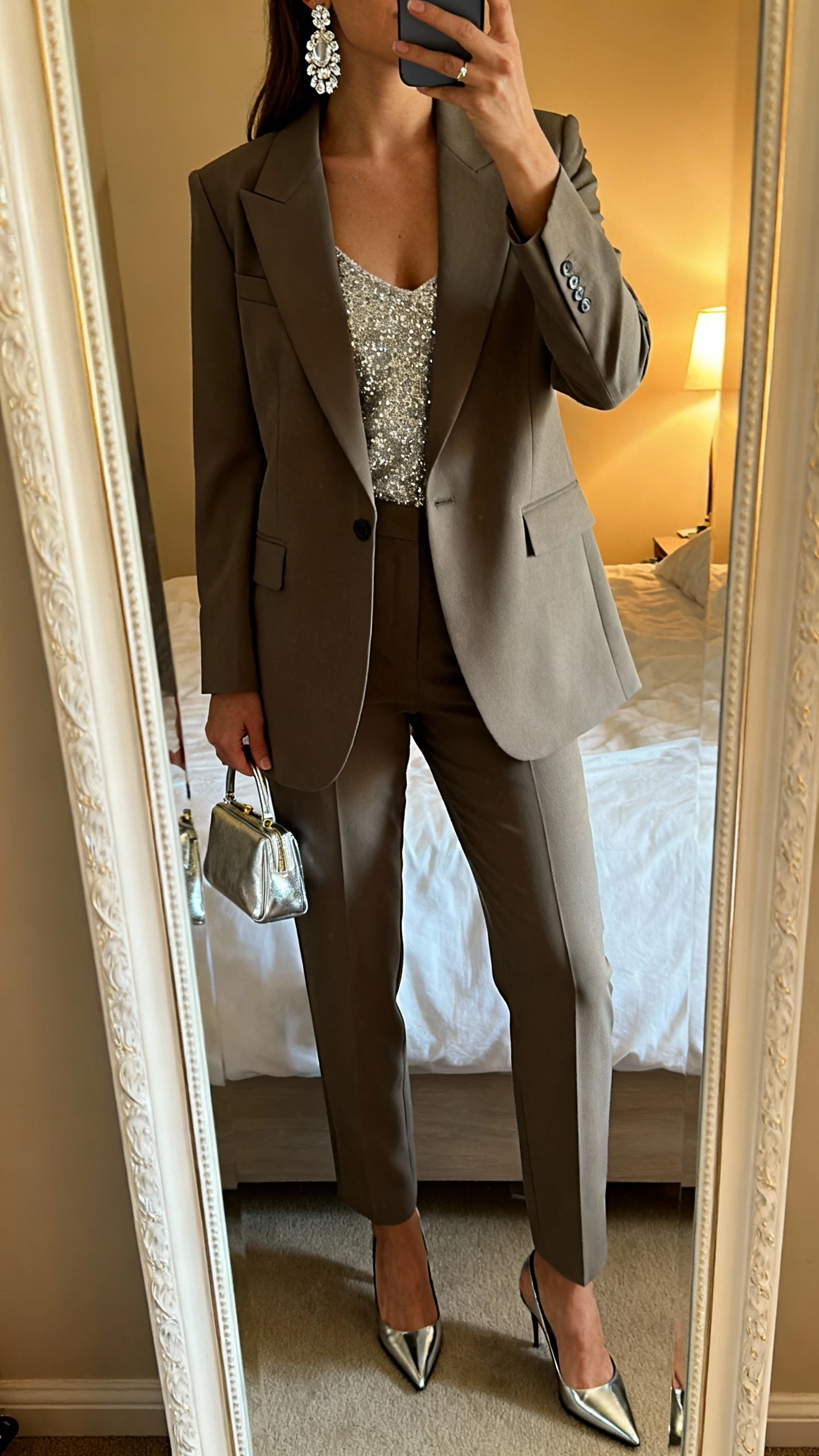 iPhone mirror selfie of a woman wearing a tailored power suit (sharp blazer and straight-leg trousers) with a sparkle camisole, statement crystal earrings, pointed-toe pumps, and a metallic mini bag, face not visible, cropped at shoulders, bedroom mirror with warm indoor lighting, iPhone photo quality.