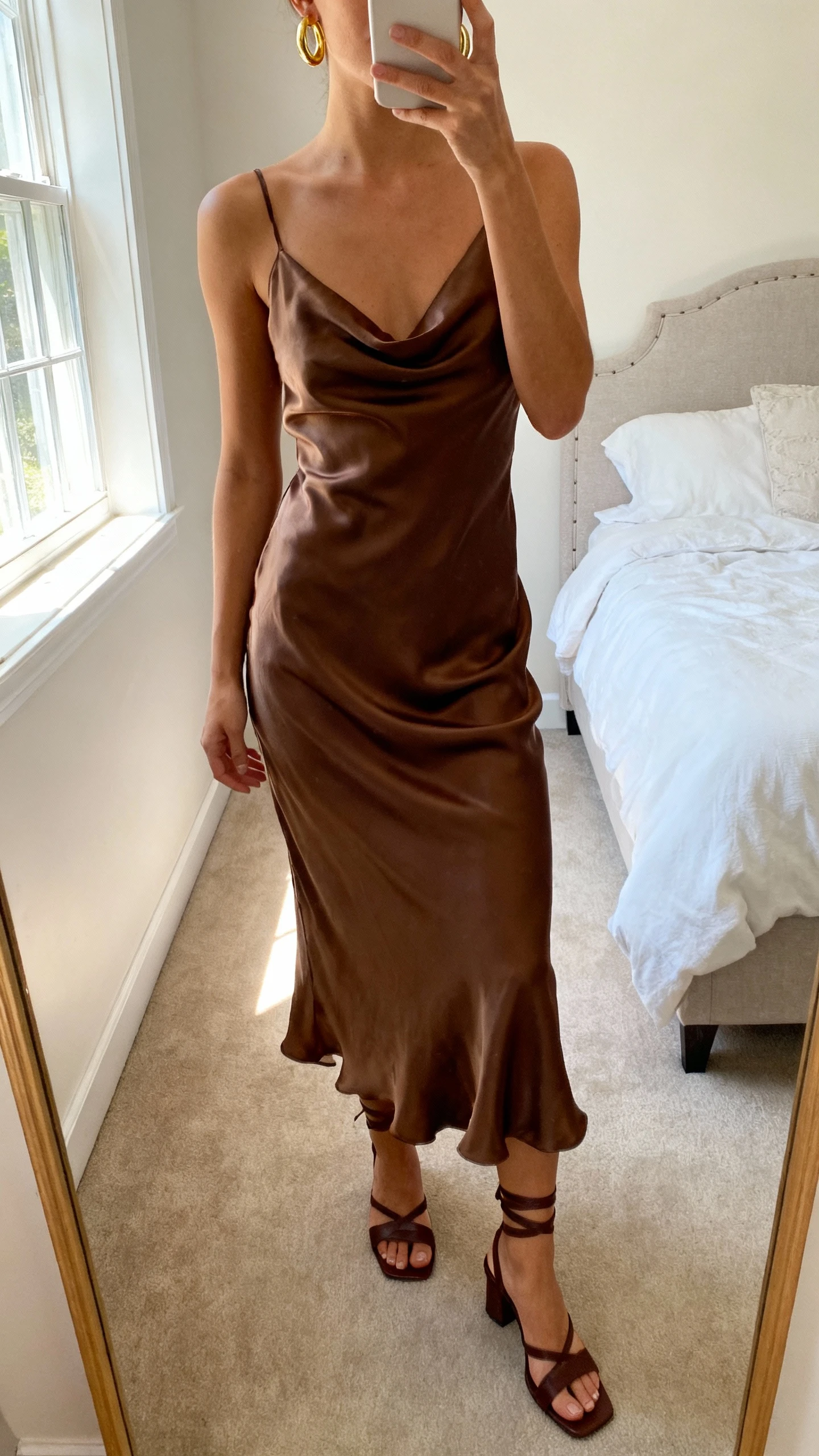 iPhone mirror selfie of a woman wearing a truffle-brown silk slip dress that drapes like ganache with delicate spaghetti straps, paired with gold hoop earrings and strappy chocolate heels, face not visible, cropped at neck, bedroom mirror, soft morning window light, iPhone photo quality.