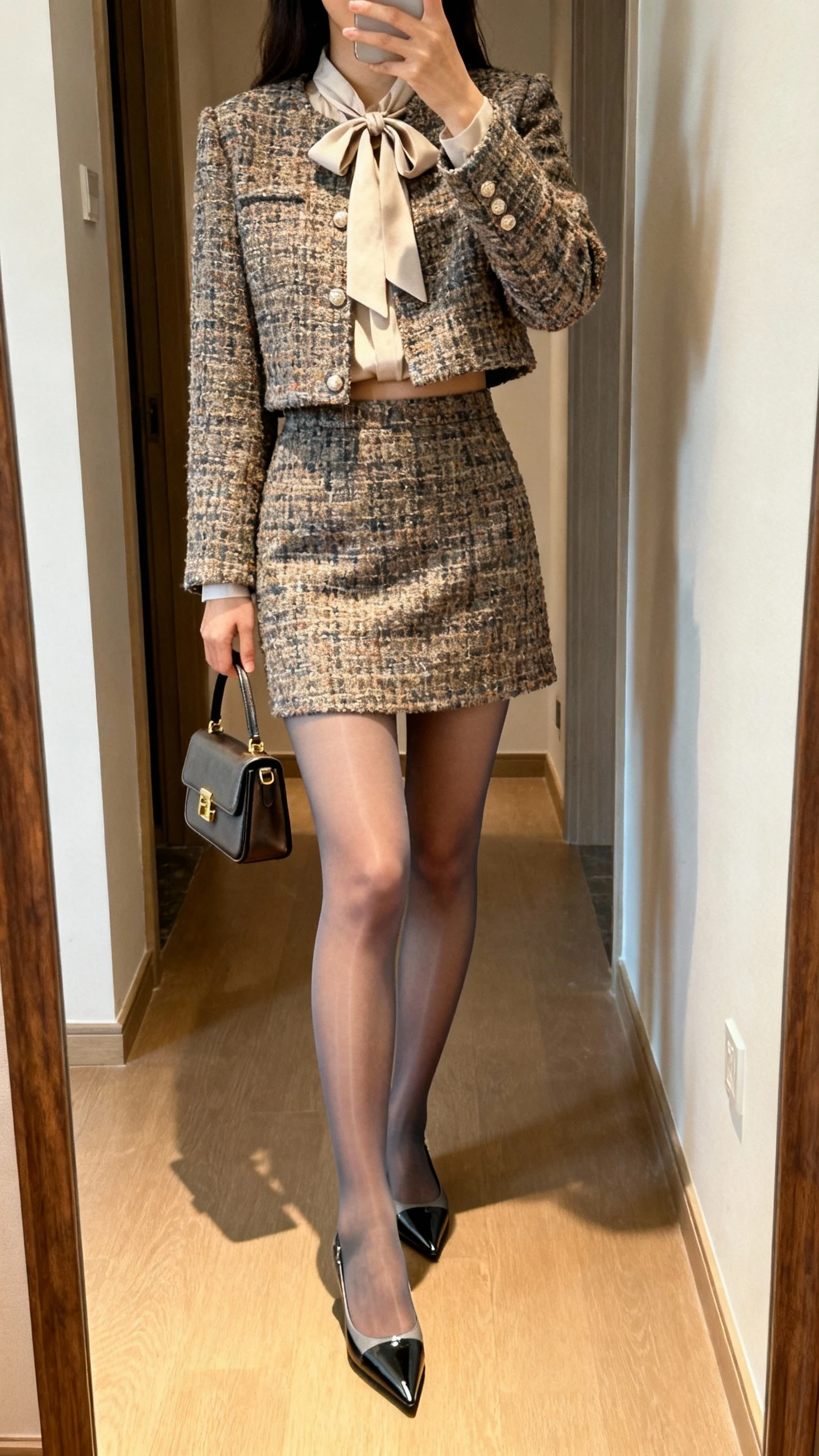iPhone mirror selfie of a woman wearing a tweed cropped jacket and mini skirt set with a bow-tie blouse, sheer tights, pointed-toe flats, and a structured mini handbag, face not visible, cropped at shoulders, full-length hallway mirror, warm indoor lighting, iPhone photo quality.