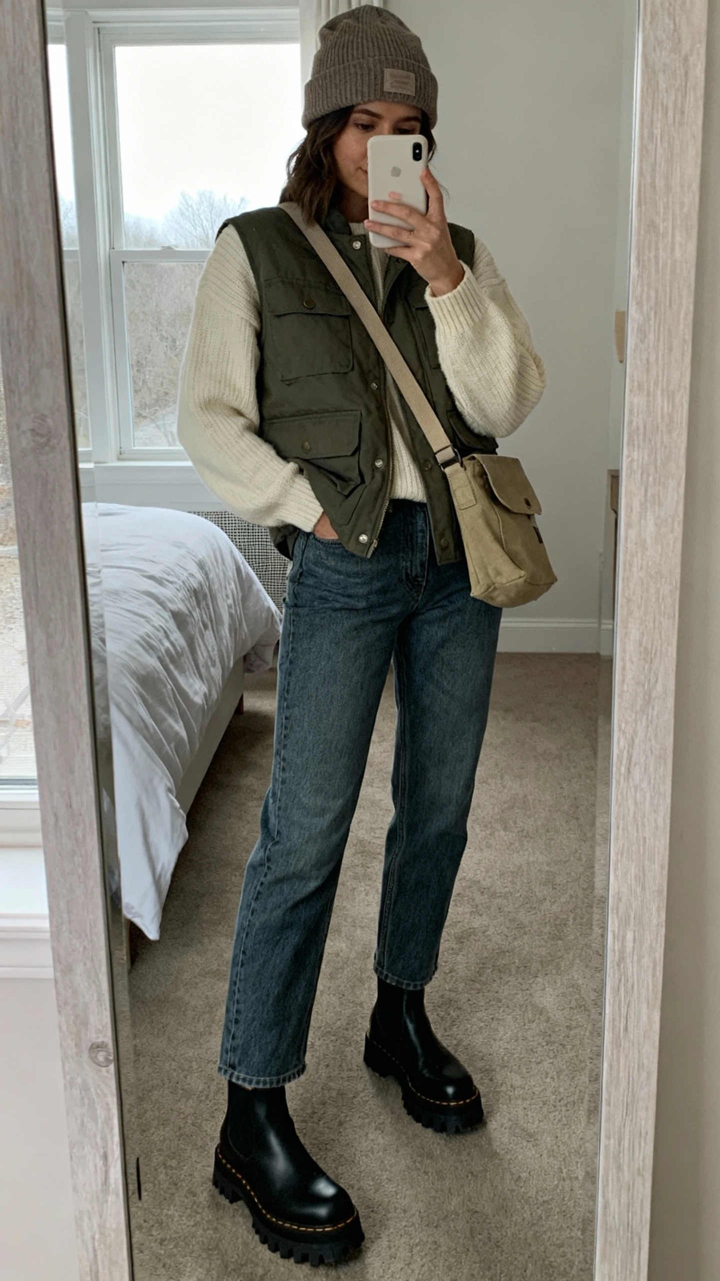 iPhone mirror selfie of a woman wearing a utility vest layered over a soft cream sweater, relaxed straight jeans, lug-sole Chelsea boots, beanie and canvas crossbody, face not visible, bedroom mirror, overcast window light, iPhone photo quality.