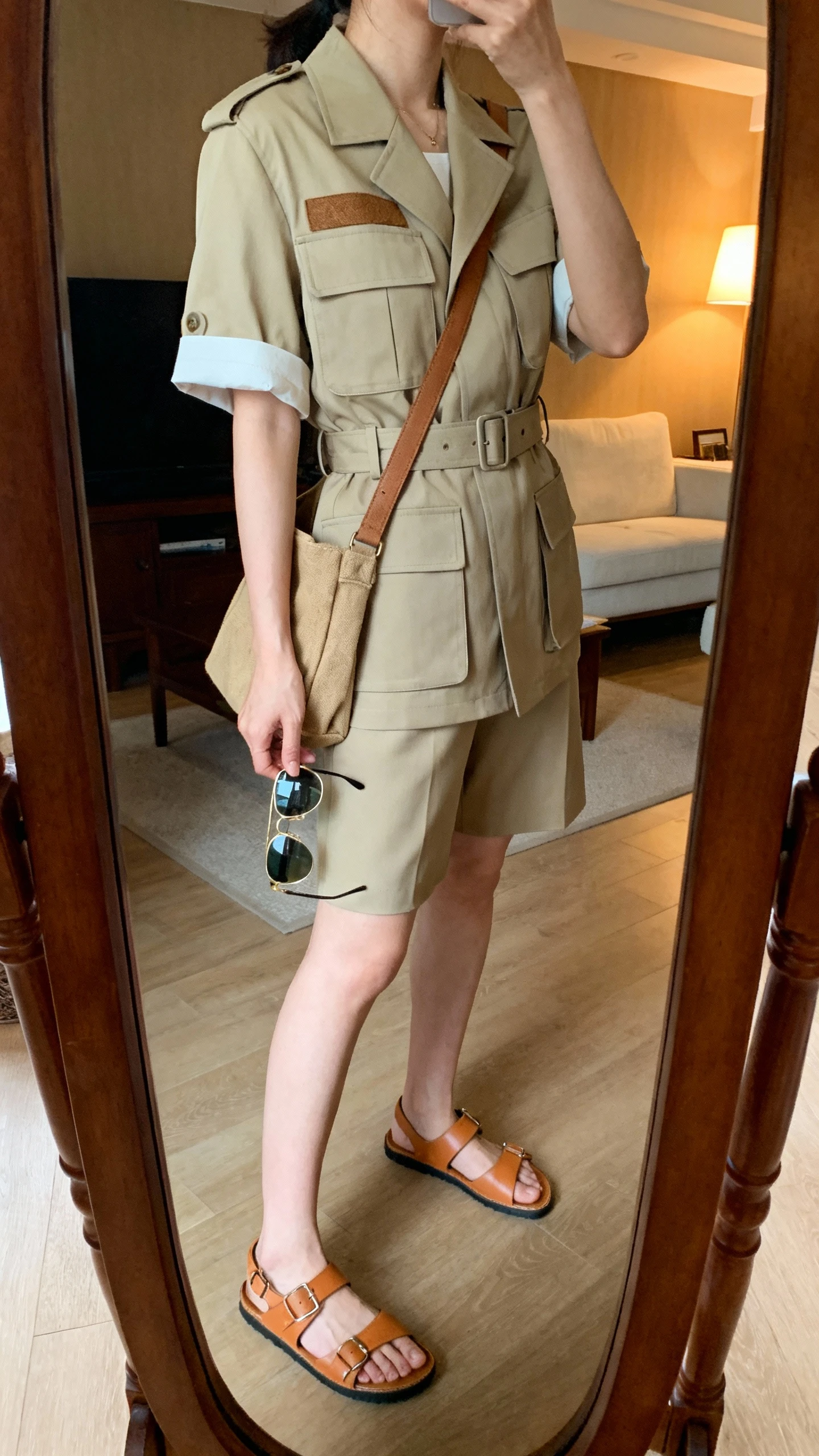 iPhone mirror selfie of a woman wearing a utility-chic safari set: short-sleeve belted utility jacket with patch pockets, matching tailored shorts, tan leather sandals, canvas crossbody, aviator-style sunglasses in hand; face not visible, side angle, living room full-length mirror with warm indoor lighting, iPhone photo quality.