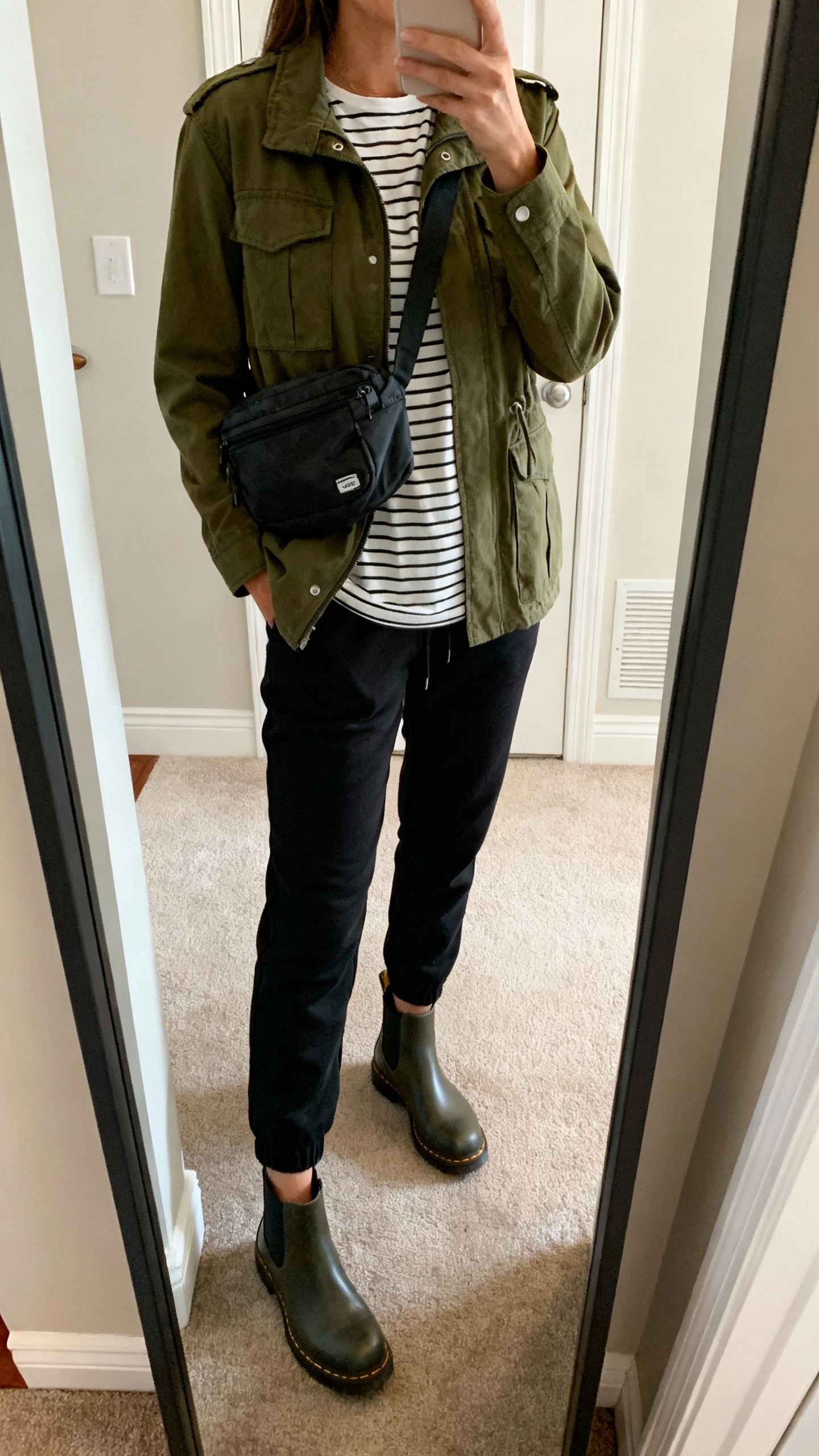iPhone mirror selfie of a woman wearing a utility jacket (army green) over a striped tee, high-rise black joggers, waterproof Chelsea boots, and a wipe-clean nylon crossbody, face not visible, cropped at shoulders, entryway mirror, soft indoor lighting, iPhone photo quality