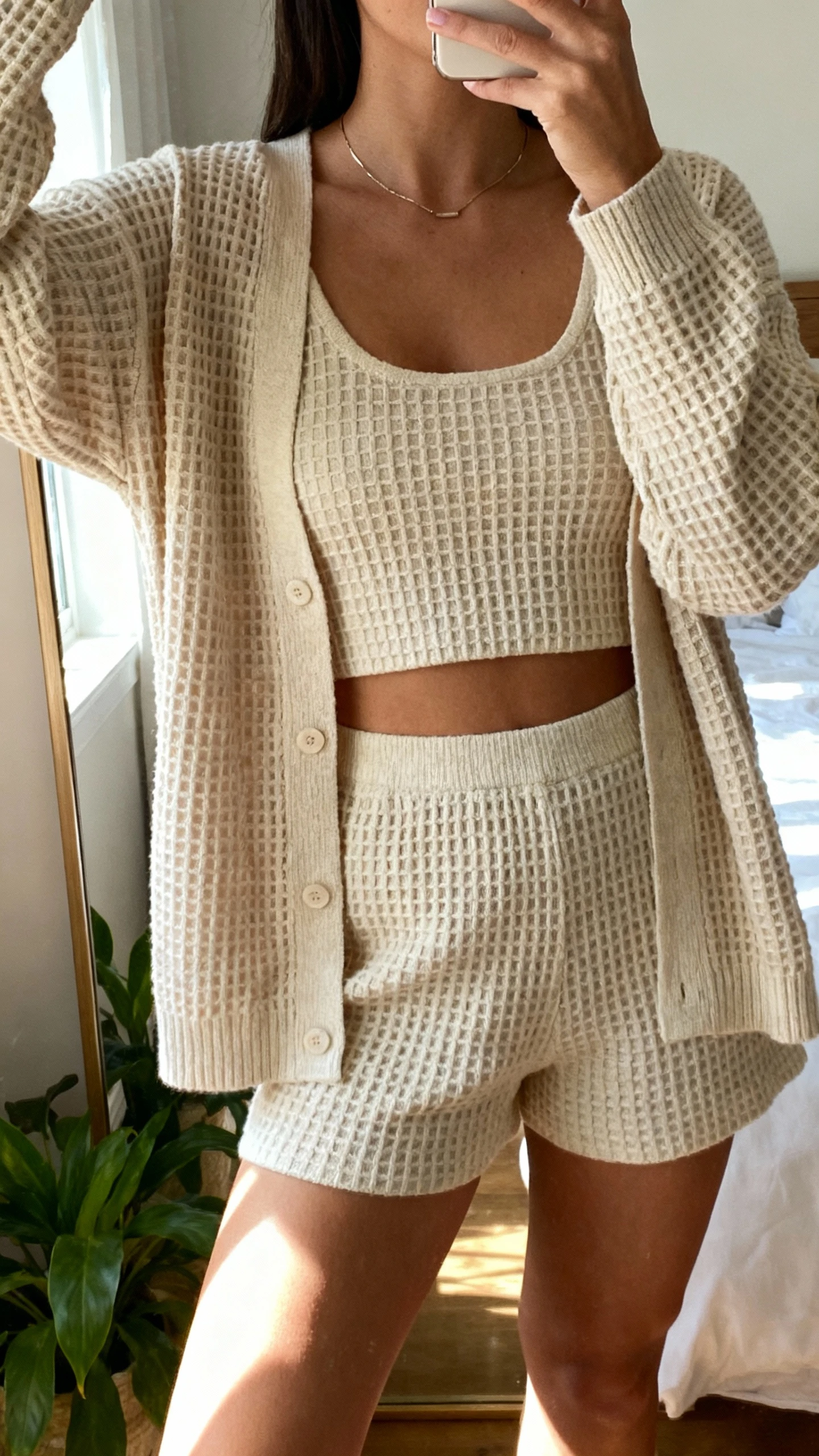 iPhone mirror selfie of a woman wearing a waffle-knit cardigan with matching knit tank and shorts, minimal necklace, face not visible, cropped at shoulders, bedroom mirror with a plant in the corner, soft afternoon light, iPhone photo quality.