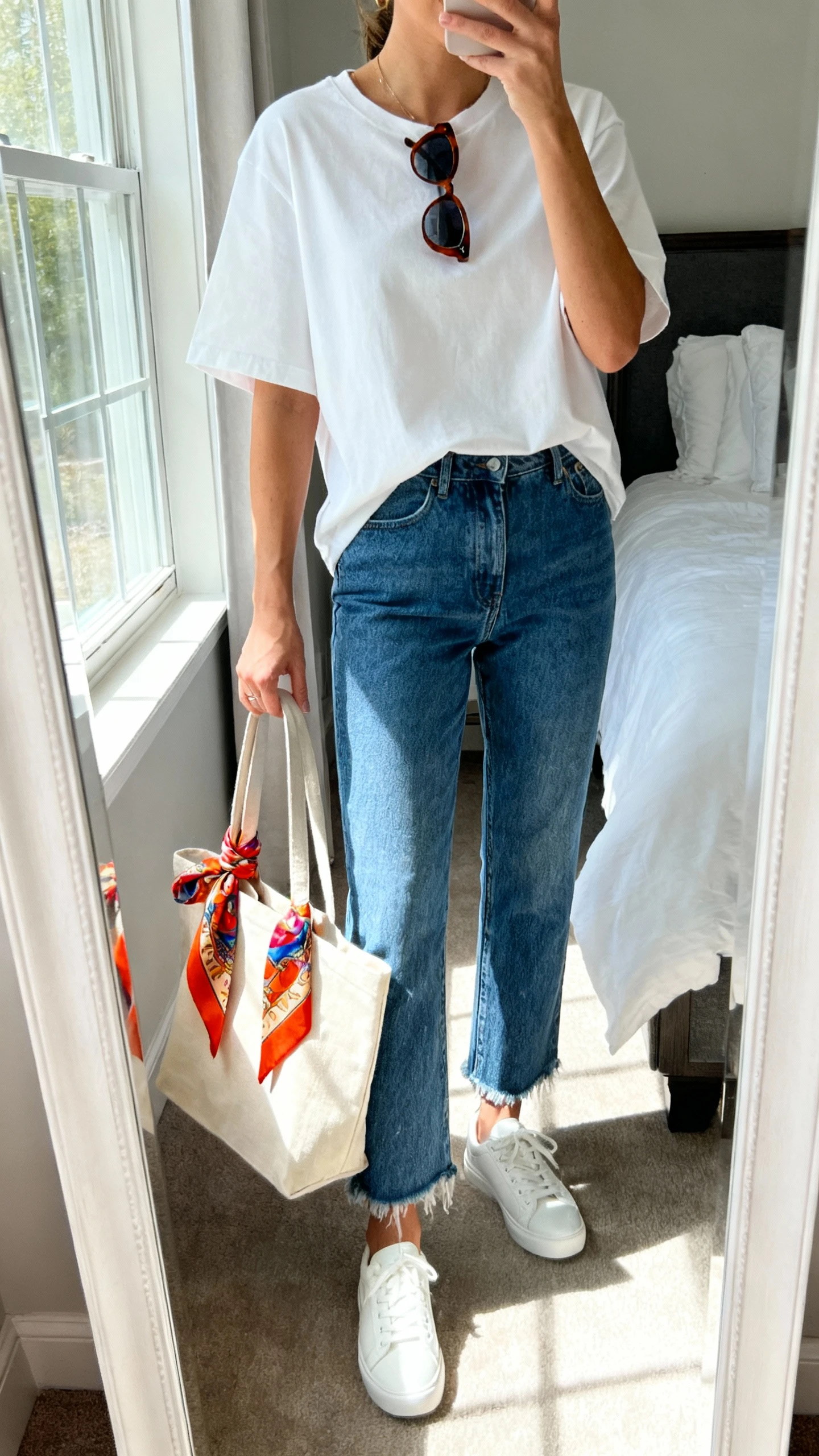 iPhone mirror selfie of a woman wearing a white boxy tee, straight-leg blue jeans with a raw hem, white sneakers, and sunglasses with a slight cat-eye, holding a canvas tote bag with a bright-print silk scarf twist-tied around one handle; face not visible, cropped at neck, bedroom mirror, morning window light, iPhone photo quality.
