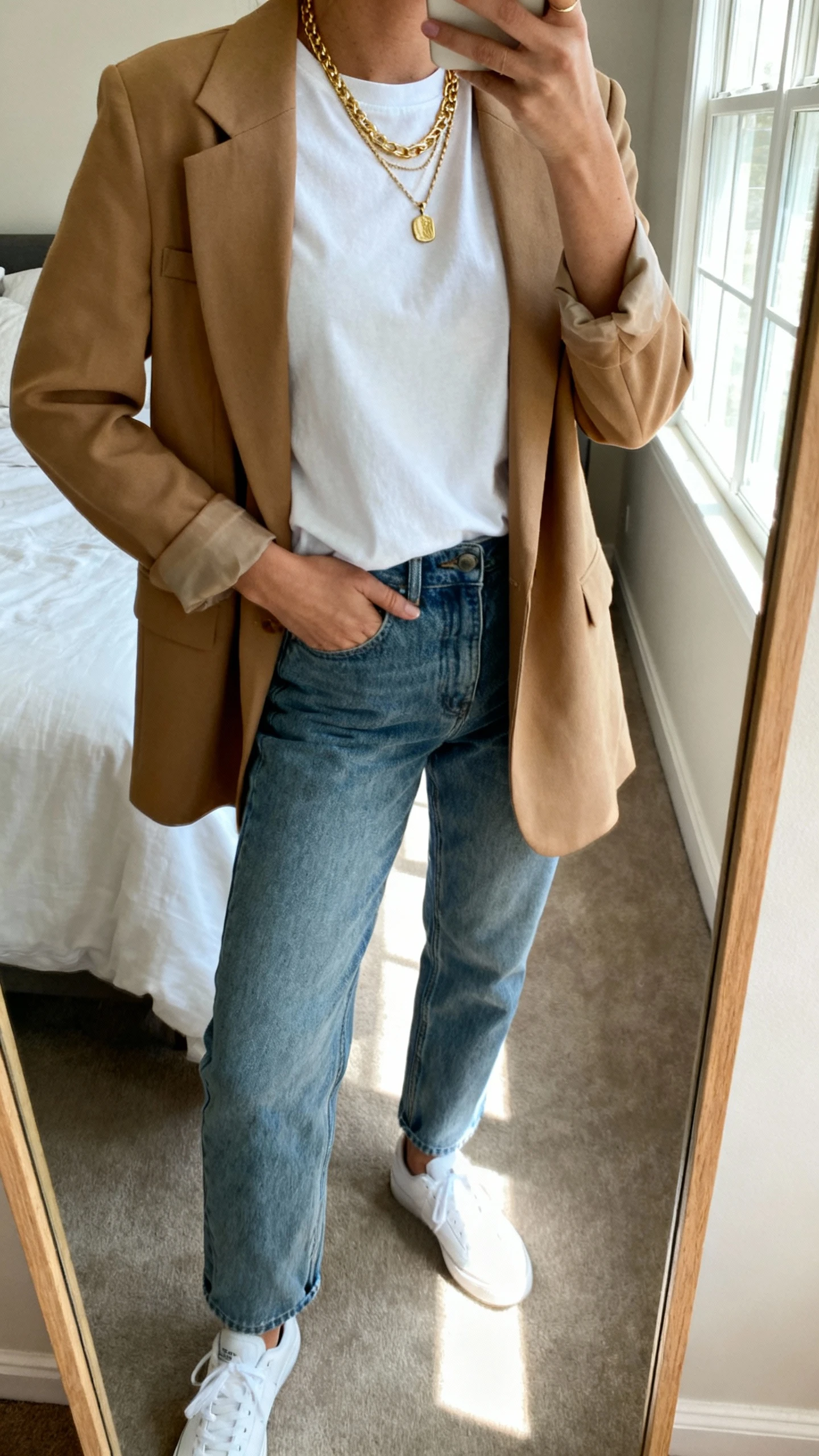 iPhone mirror selfie of a woman wearing a white crewneck tee, relaxed jeans, a camel blazer, white sneakers, and layered gold necklaces (short chain + pendant + longer chain), face not visible, cropped at neck, bedroom mirror with natural window light, iPhone photo quality.