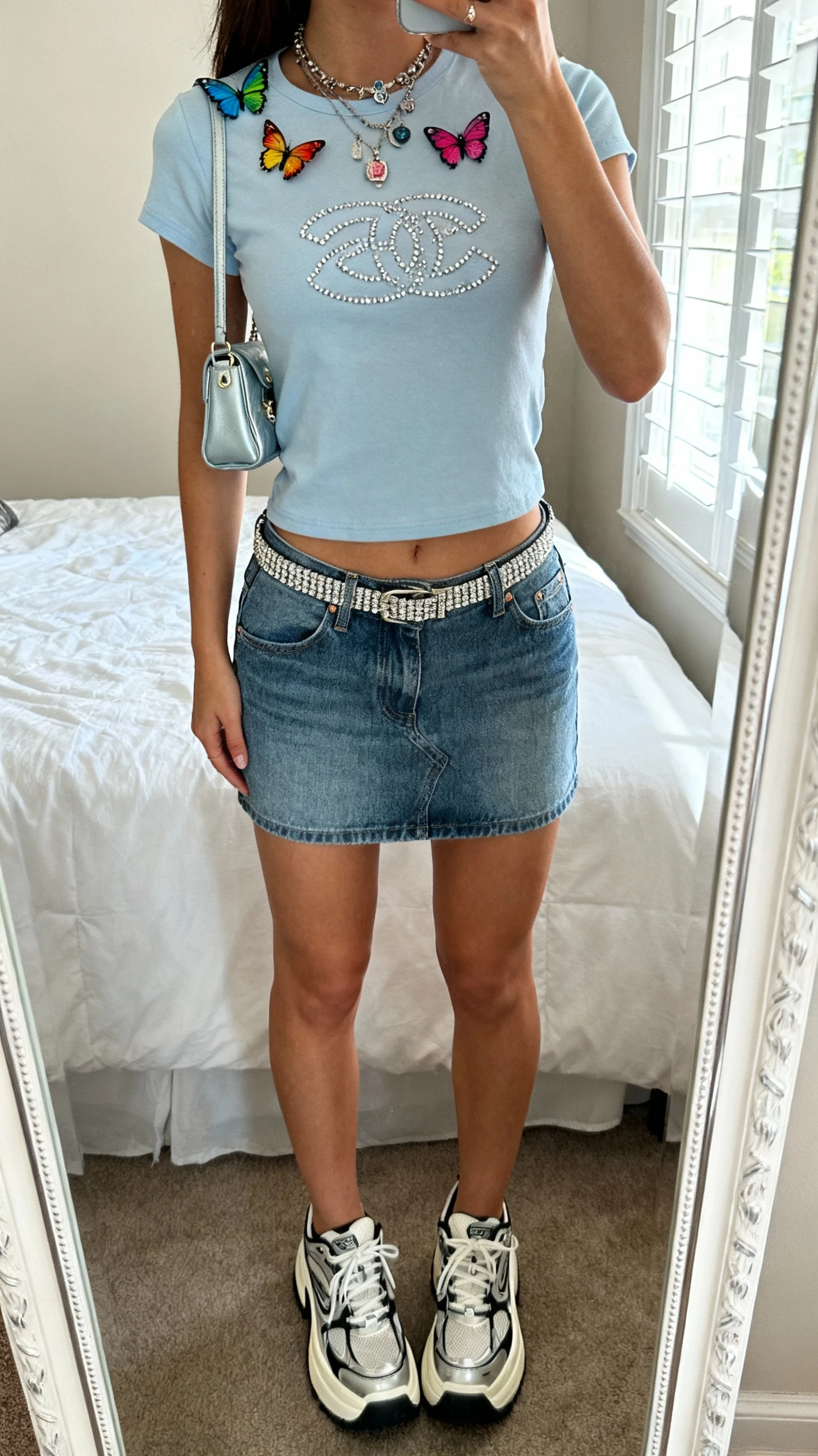 iPhone mirror selfie of a woman wearing a Y2K low-rise denim mini skirt, baby tee with a rhinestone graphic, chunky platform sneakers, thin rhinestone belt, tiny shoulder baguette bag, colorful butterfly hair clips, and layered charm necklace, face not visible, cropped at neck, bedroom mirror, natural window light, iPhone photo quality