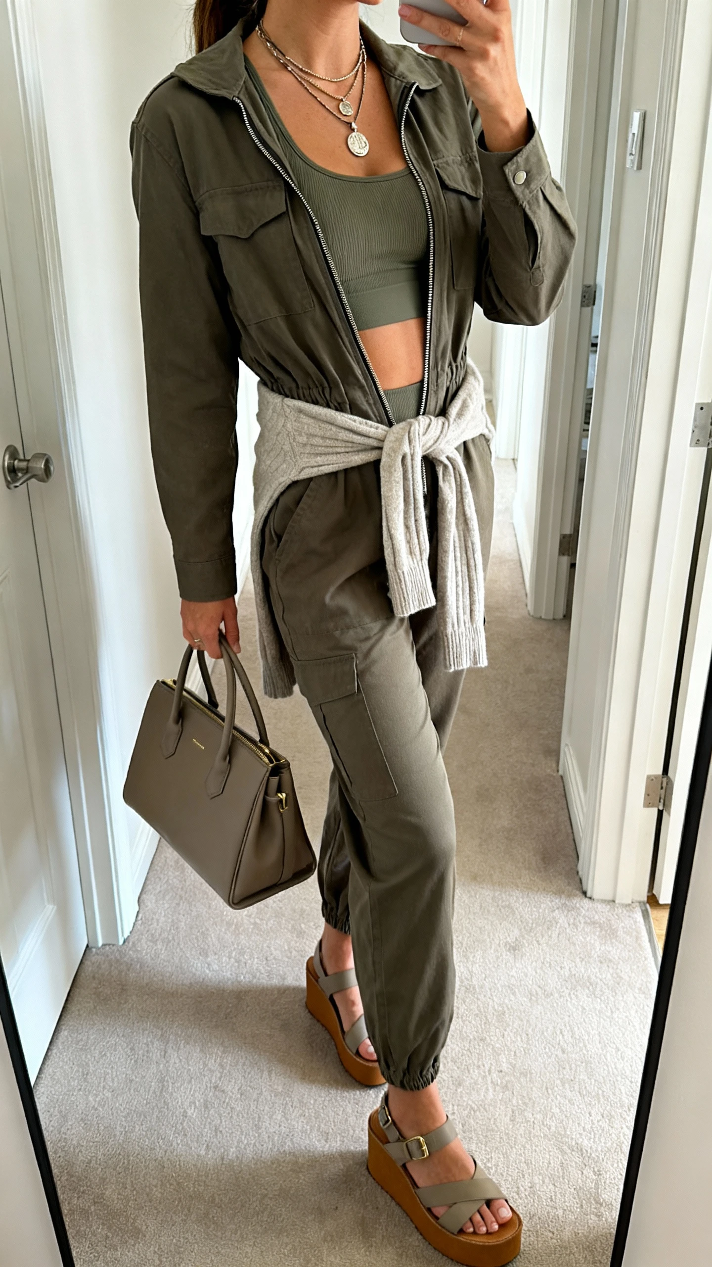 iPhone mirror selfie of a woman wearing a zip-front utility jumpsuit layered over a fitted sports bra, unzipped slightly at top, tied sweater at waist, platform sandals, layered pendant necklace, structured tote, face not visible, side angle in hallway mirror with natural daylight, iPhone photo quality.