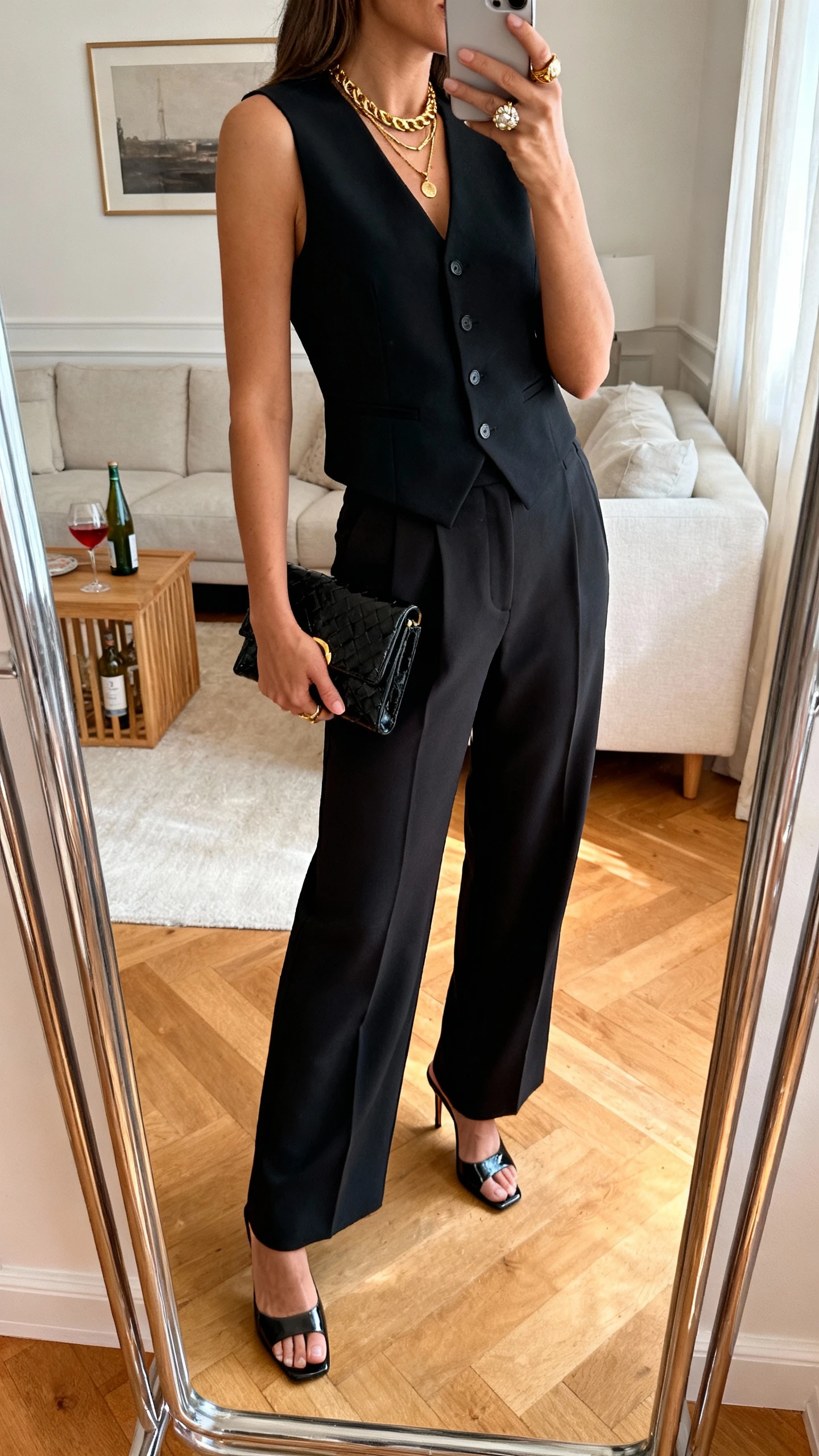 iPhone mirror selfie of a woman wearing an aperitivo-to-night out capsule: black tailored vest top, high-waist wide-leg trousers, sleek kitten-heel mules, compact clutch, layered gold necklaces, and statement ring; face not visible, side angle in living room mirror with warm indoor lighting, iPhone photo quality.