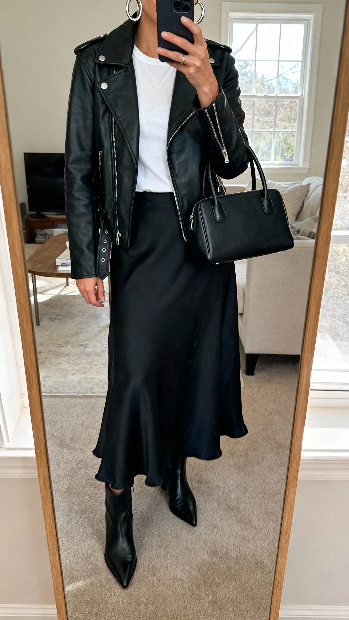 iPhone mirror selfie of a woman wearing an effortless maxi skirt and moto jacket: black leather moto jacket, white fitted tee, flowy black maxi skirt, pointed black ankle boots, silver hoops, black top-handle bag, face not visible, cropped at neck, living room full-length mirror, natural window light, iPhone photo quality