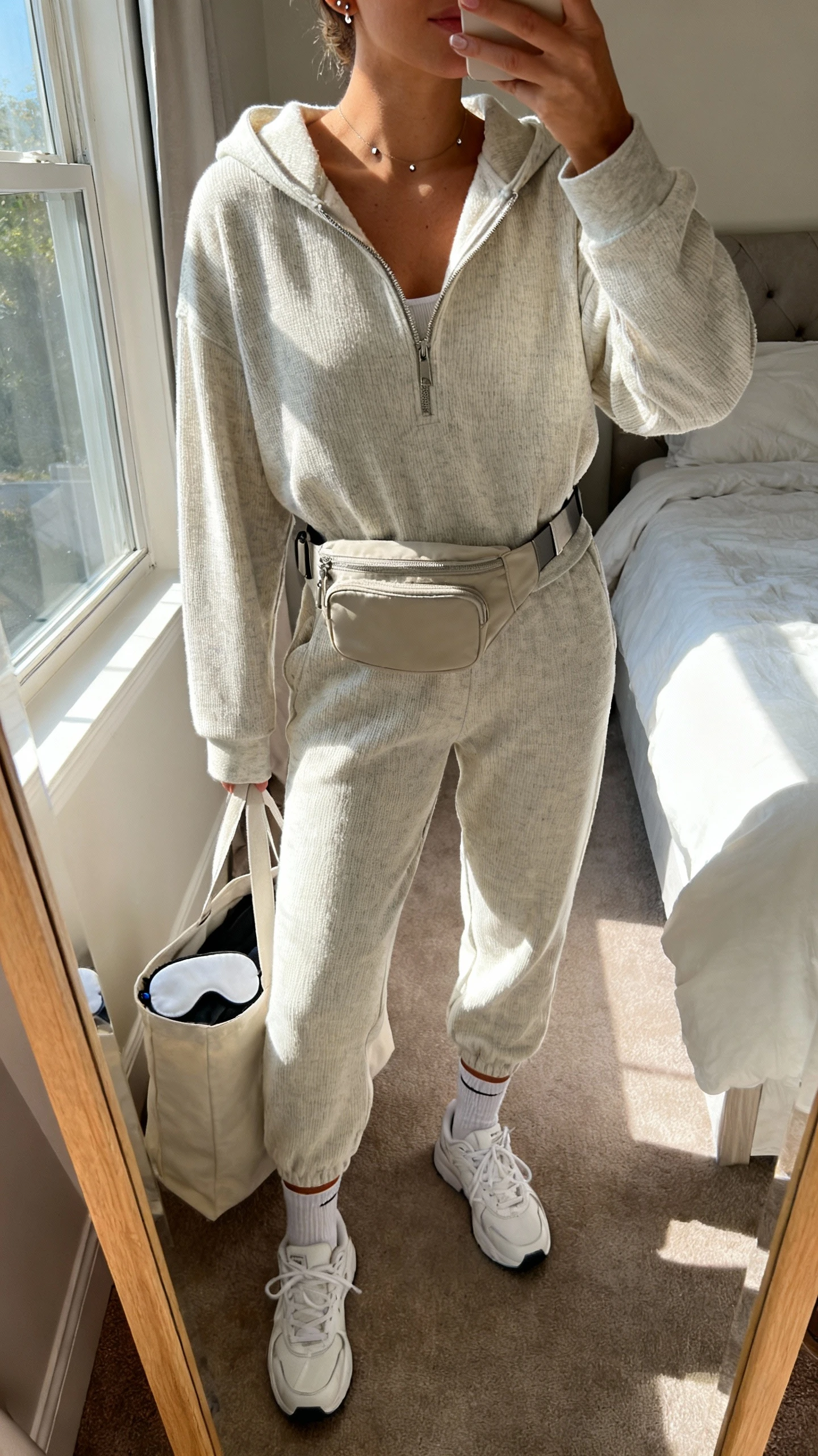 iPhone mirror selfie of a woman wearing an elevated matching jogger set (soft knit joggers with a coordinated zip hoodie), sleek white sneakers, crossbody belt bag, thin crew socks, minimalist studs, and an eye mask peeking from tote; face not visible, cropped at neck, bedroom mirror with morning window light, iPhone photo quality.