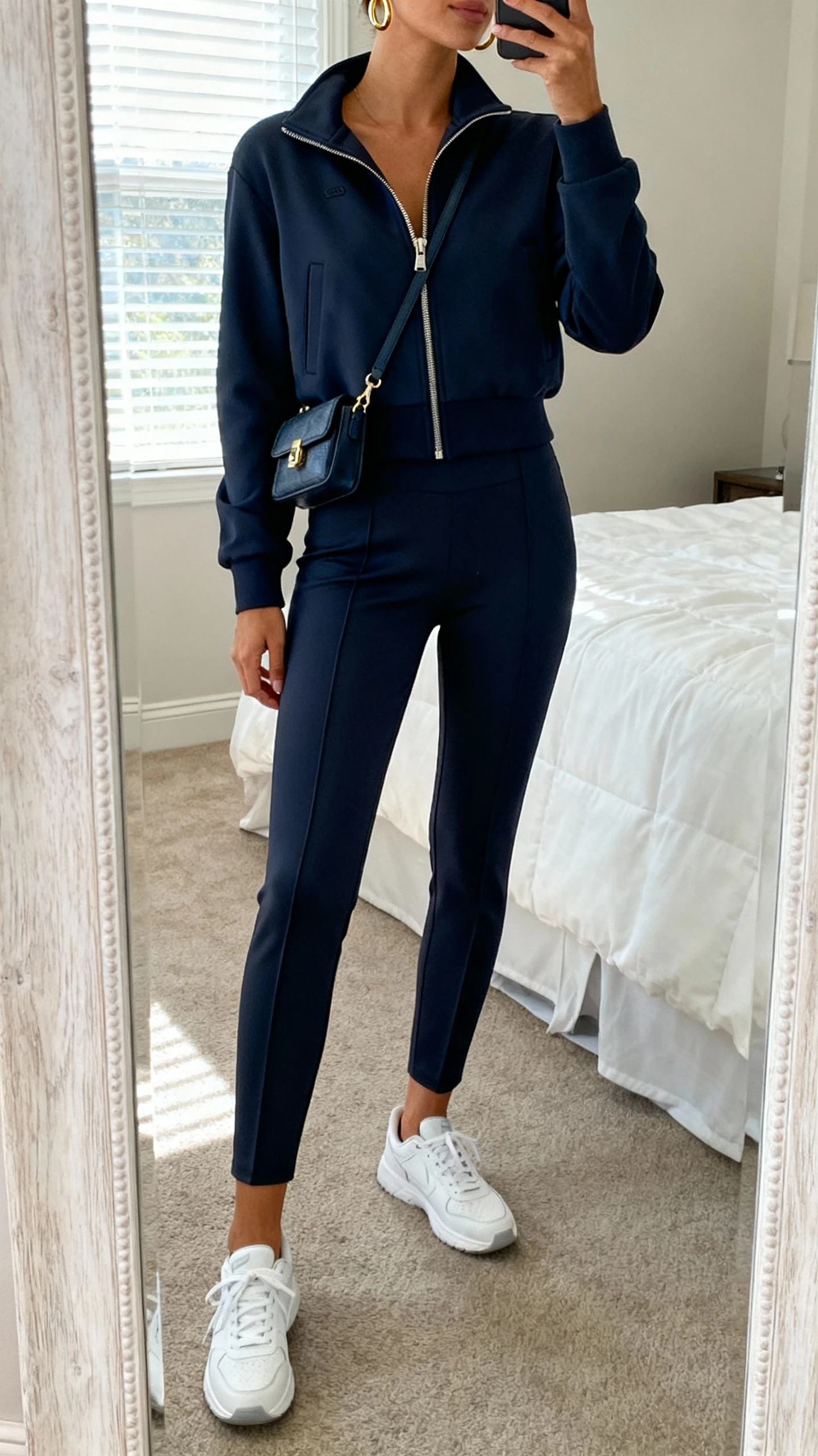 iPhone mirror selfie of a woman wearing an elevated athleisure set: matching tailored leggings and zip-up track jacket with clean white sneakers, small crossbody bag, minimal gold hoops, face not visible, cropped at neck, bedroom mirror with natural window light, iPhone photo quality.