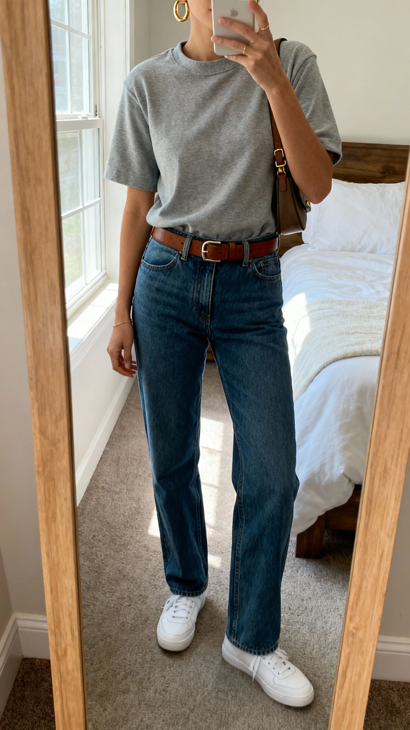 iPhone mirror selfie of a woman wearing an elevated crewneck tee tucked into high-rise straight-leg jeans with a leather belt, minimalist white sneakers, delicate gold hoop earrings, and a crossbody bag, face not visible, cropped at neck, bedroom mirror, natural window light, iPhone photo quality.