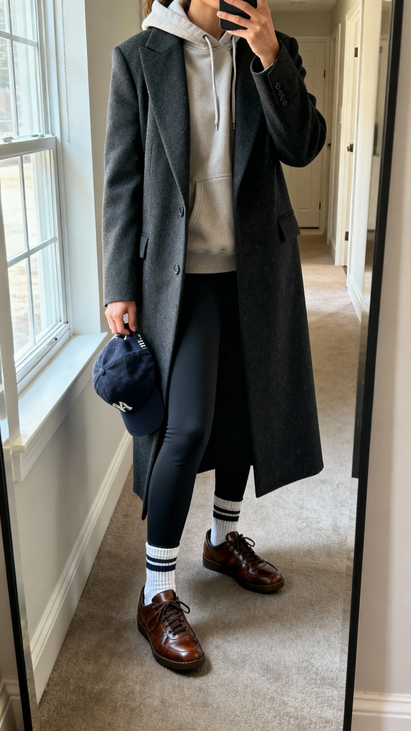 iPhone mirror selfie of a woman wearing an elevated hoodie layered under a tailored long coat with straight-leg leggings, sporty crew socks, clean leather sneakers, and a baseball cap in hand, face not visible, cropped at neck, hallway full-length mirror, natural window light, iPhone photo quality.