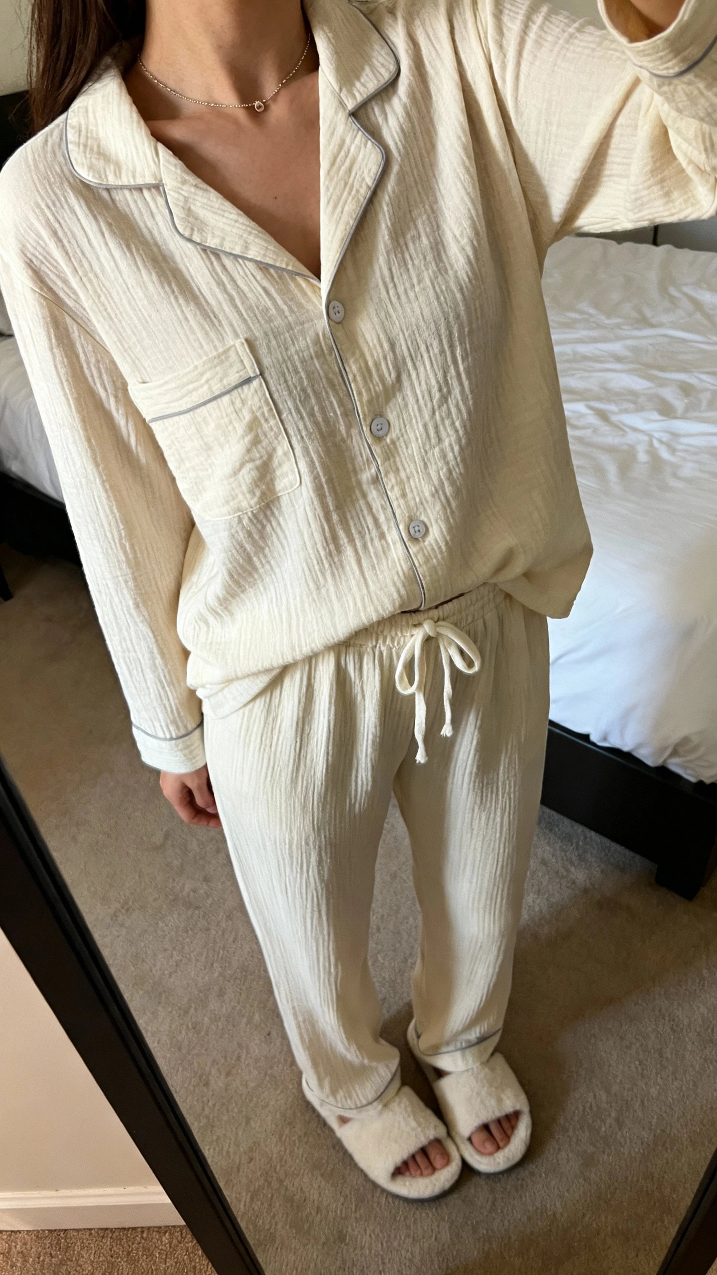 iPhone mirror selfie of a woman wearing an elevated pajama set (button-up top and matching drawstring pants) with house slippers and a delicate necklace, face not visible, cropped at shoulders, bedroom mirror, warm indoor lighting, iPhone photo quality.