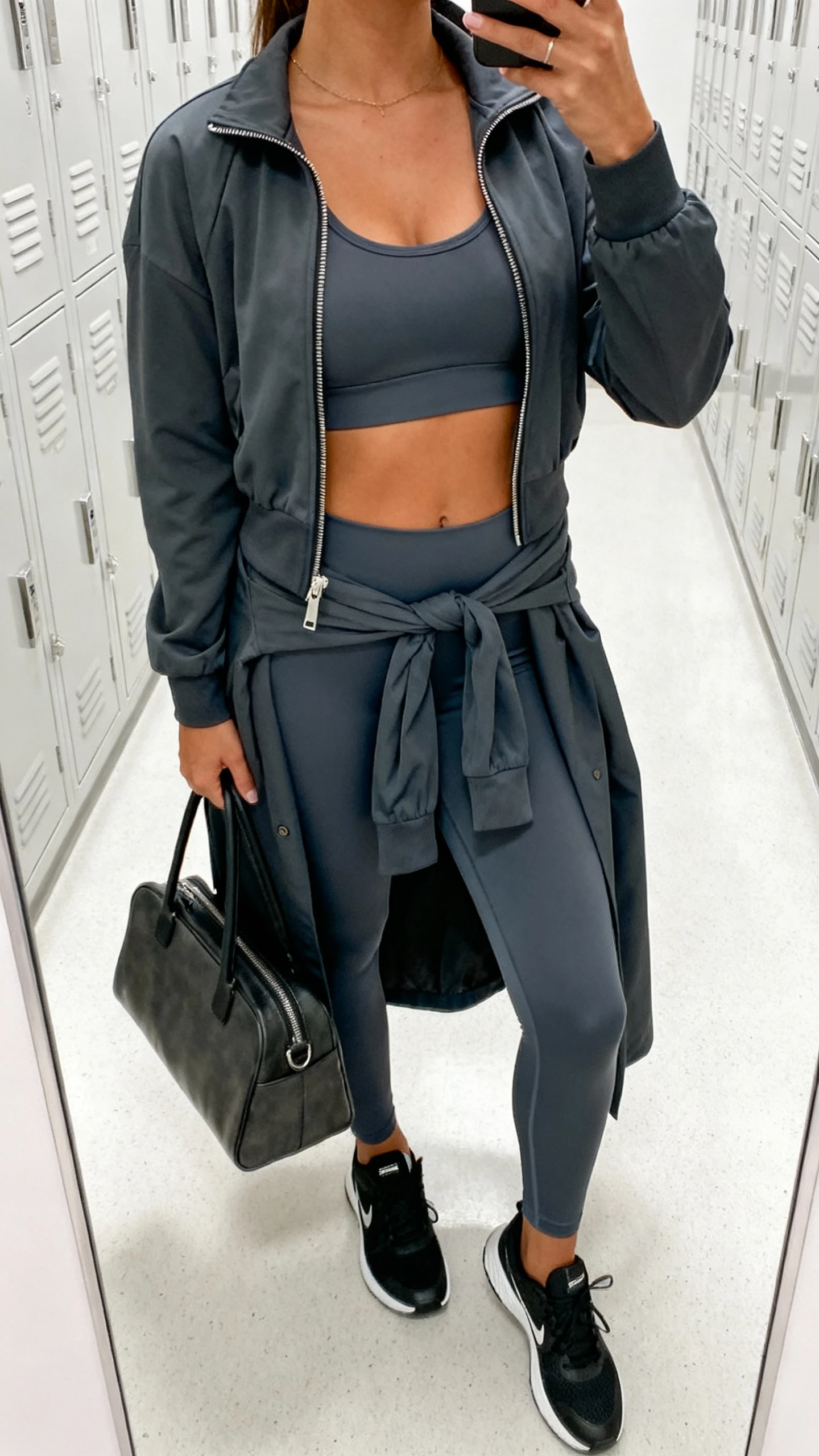 iPhone mirror selfie of a woman wearing an elevated athleisure set: matching high-waist leggings and zip-front track jacket over a sports bra, layered with a longline coat tied at the waist, sleek trainers, and a structured tote; face not visible, cropped at neck, gym locker-room mirror with bright indoor lighting, iPhone photo quality.