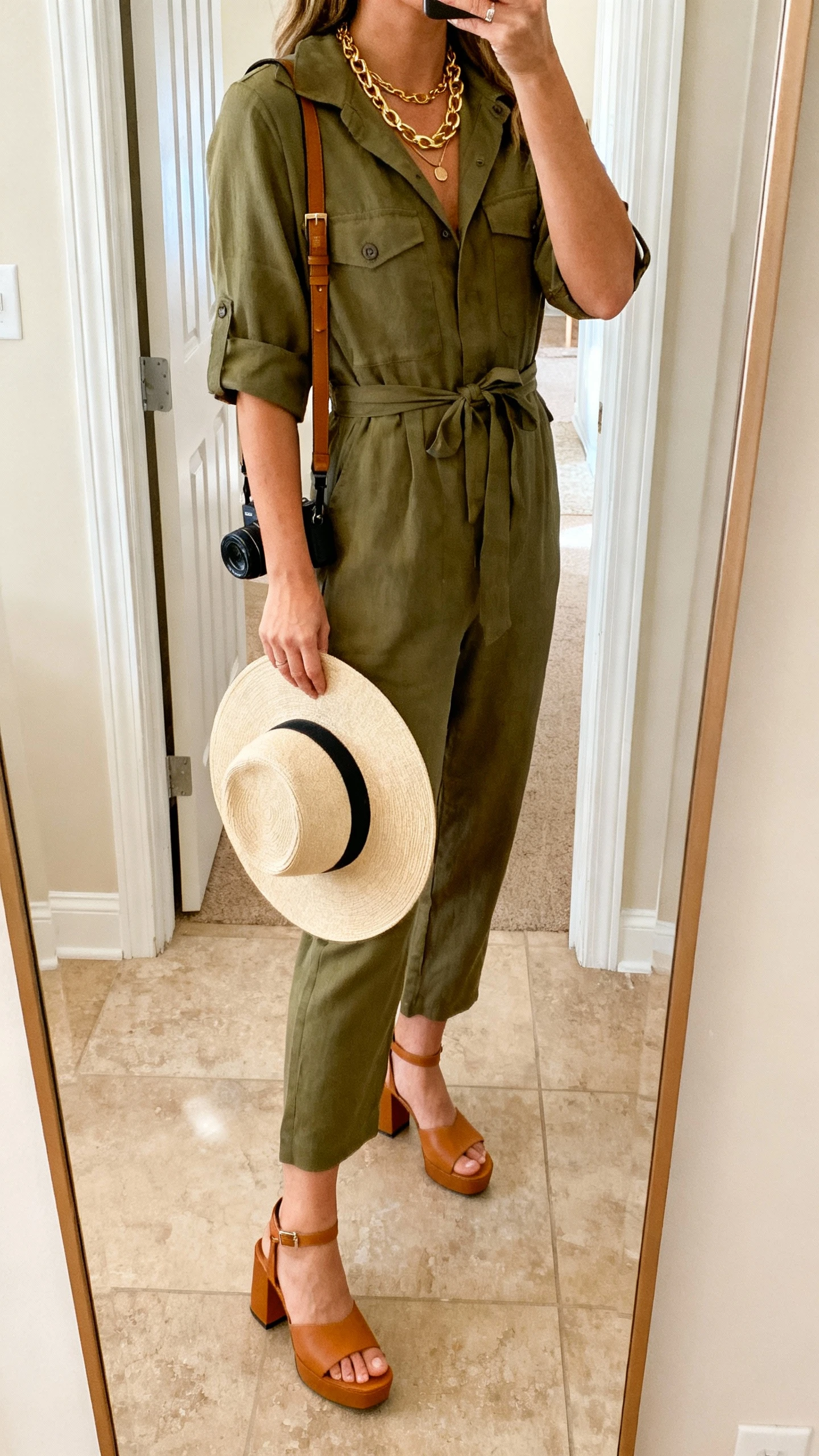 iPhone mirror selfie of a woman wearing an olive utility jumpsuit cinched at the waist, tan block-heel sandals, layered gold necklaces, a wide-brim hat in hand, and a crossbody camera bag, face not visible, side angle, entryway mirror, warm indoor lighting, iPhone photo quality