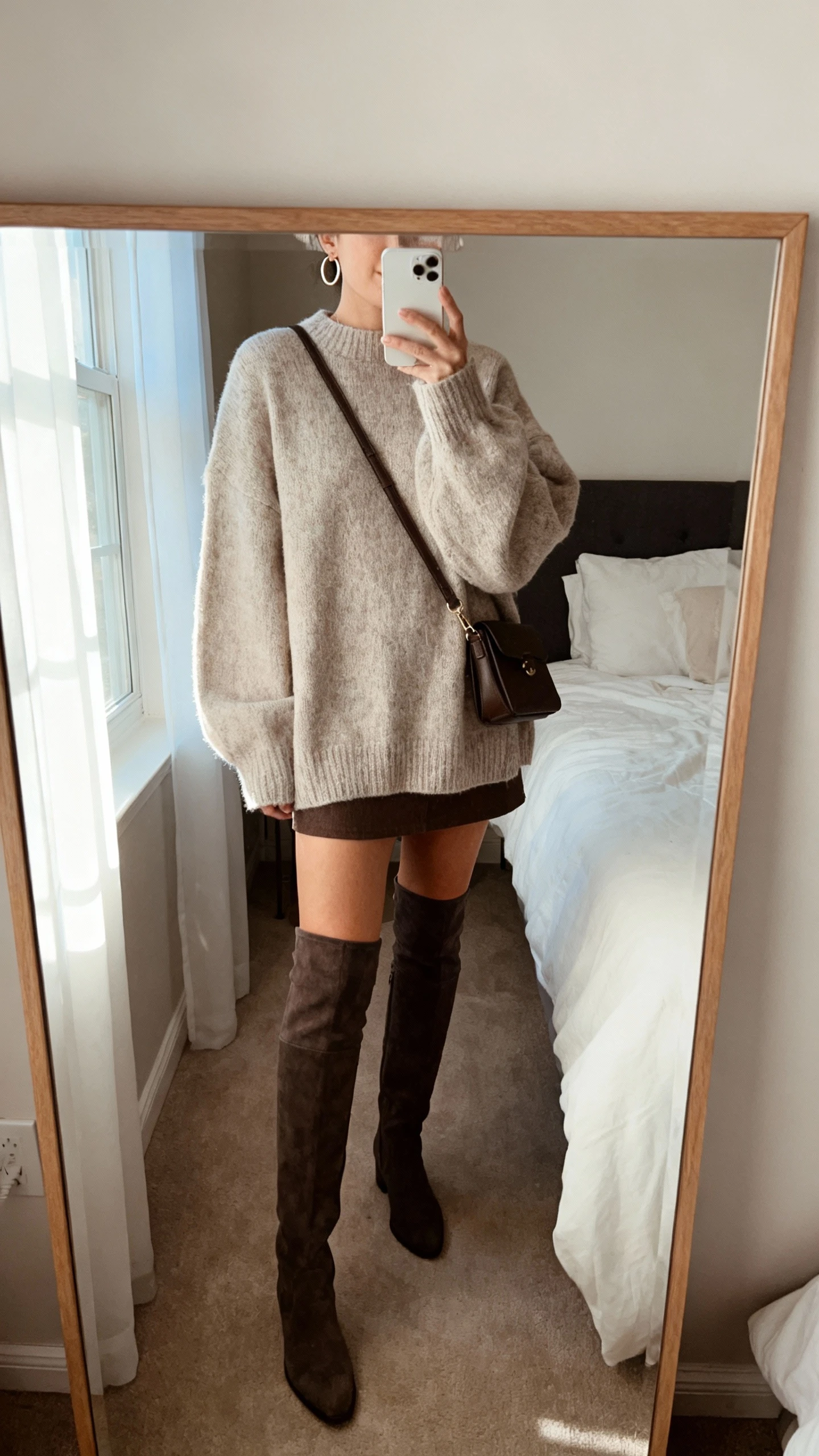iPhone mirror selfie of a woman wearing an oversized cozy sweater, a mini skirt, and over-the-knee boots, face not visible, cropped at neck, bedroom mirror with soft morning window light, iPhone photo quality; simple crossbody bag and minimal hoop earrings.