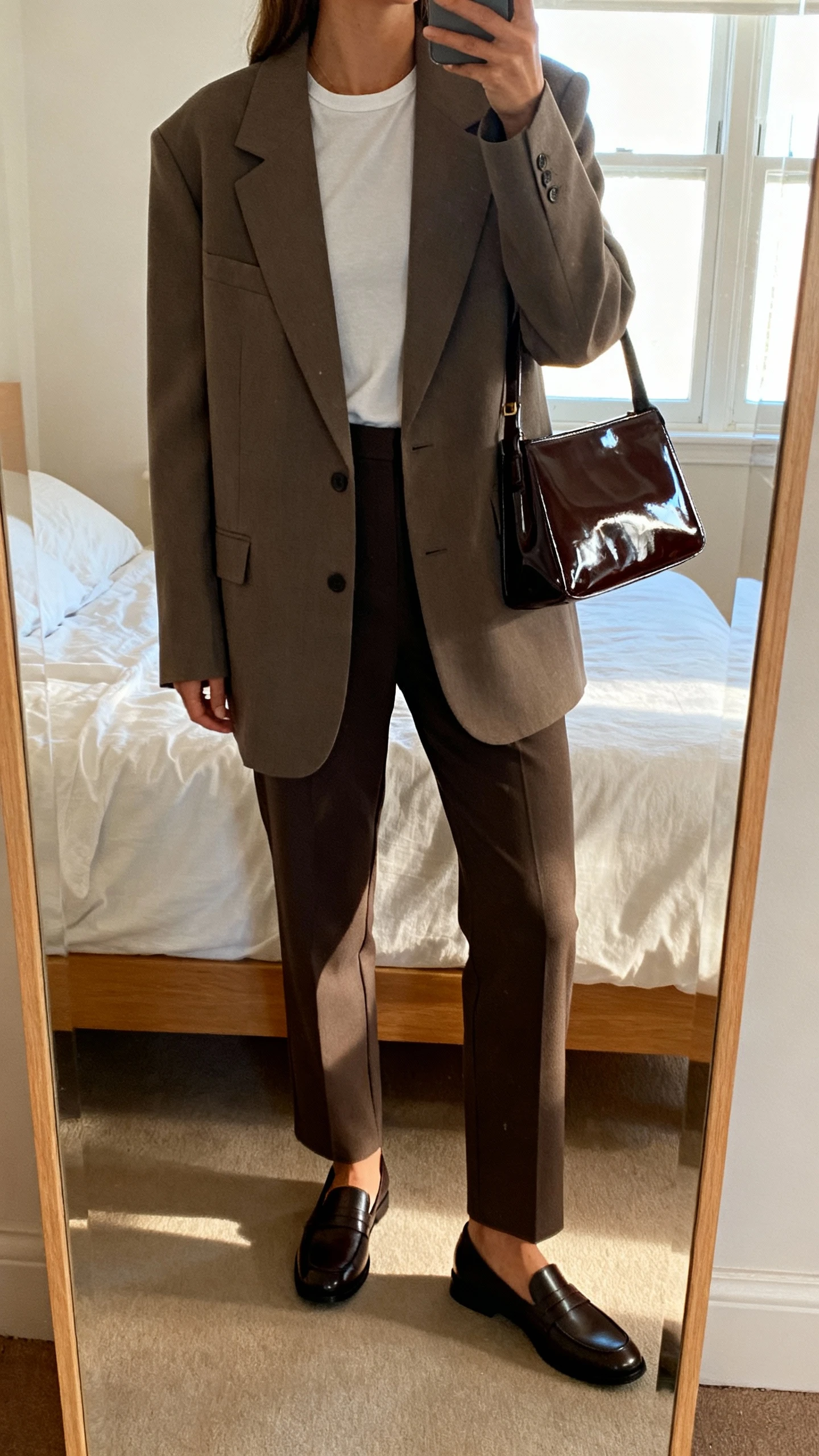 iPhone mirror selfie of a woman wearing an oversized power blazer over a minimalist tee, tailored straight-leg trousers, sleek loafers, and a high-gloss patent shoulder bag; face not visible, cropped at neck, bedroom mirror, morning window light, iPhone photo quality.