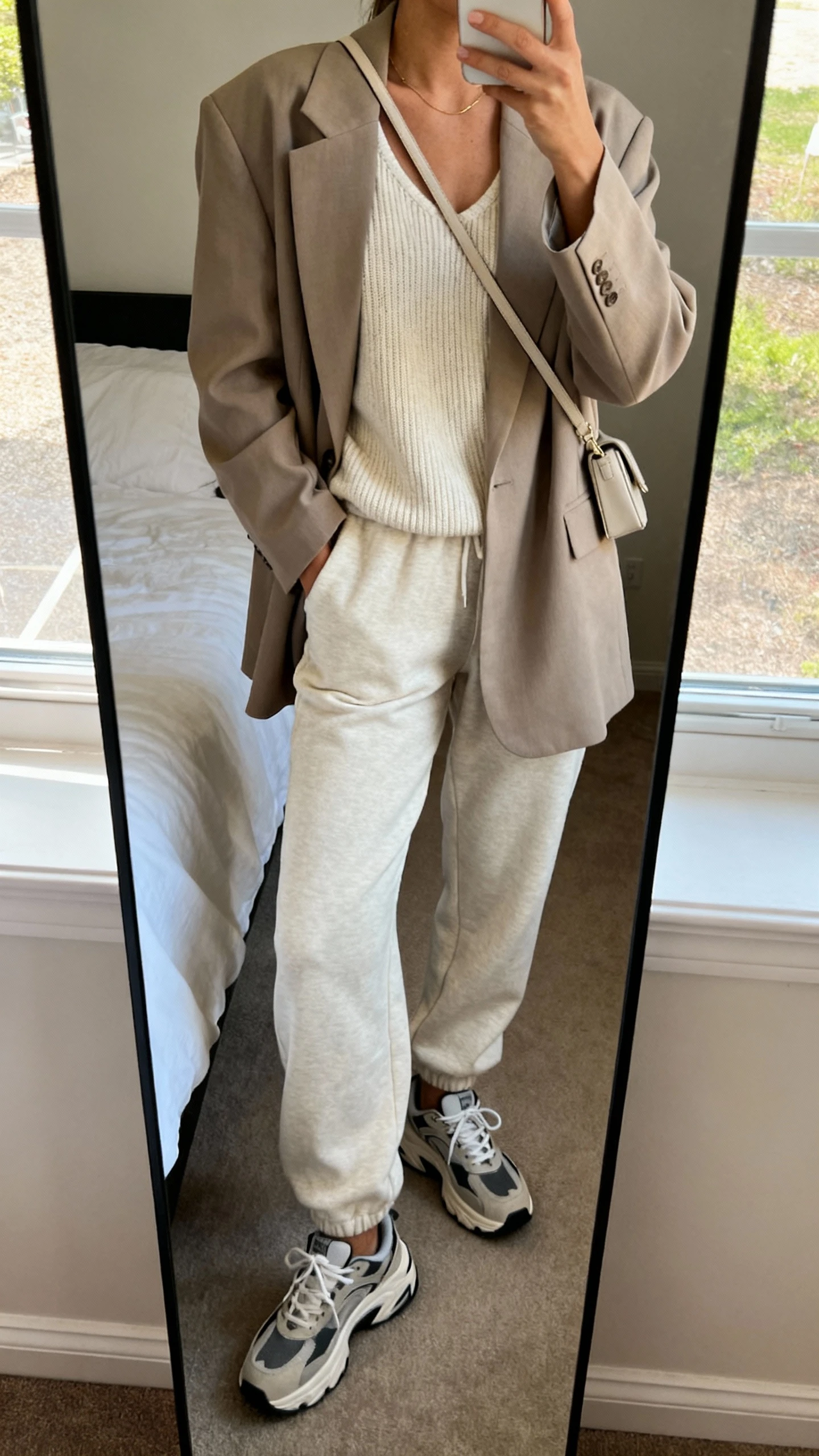 iPhone mirror selfie of a woman wearing an oversized blazer layered over a matching lounge set (soft knit top and relaxed joggers), chunky sneakers, minimalist crossbody, face not visible, cropped at neck, bedroom full-length mirror, natural window light, iPhone photo quality.