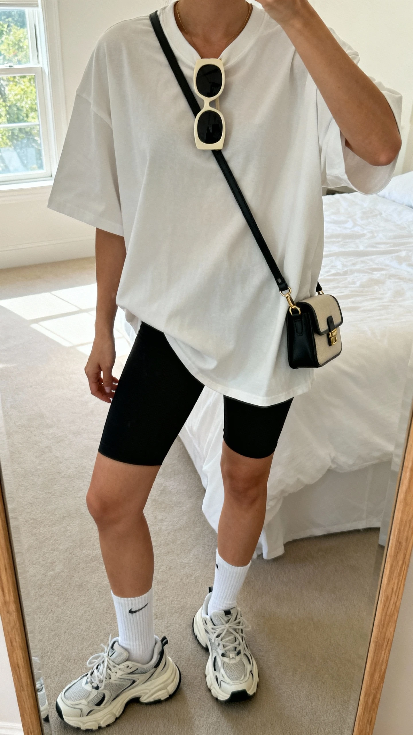 iPhone mirror selfie of a woman wearing an oversized T-shirt with black bike shorts, oversized sunglasses, white crew socks, chunky dad sneakers, and a small crossbody bag, face not visible, cropped at neck, bedroom mirror with natural window light, iPhone photo quality.