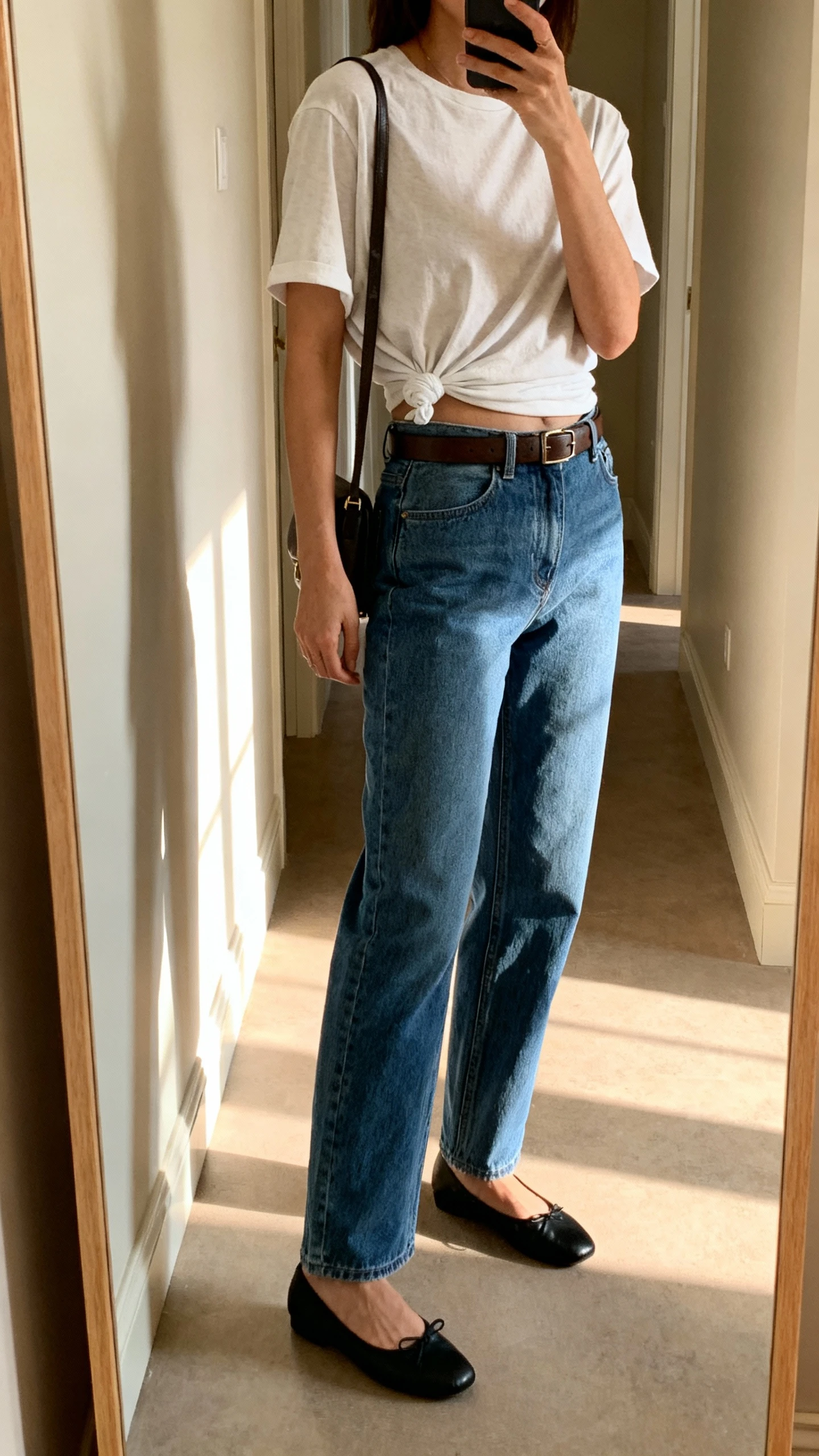 iPhone mirror selfie of a woman wearing an oversized T-shirt half-tucked (French tuck) into straight-leg blue denim, classic black ballet flats, a slim leather belt, and a simple shoulder bag, face not visible, side angle in a hallway mirror with morning light, iPhone photo quality.
