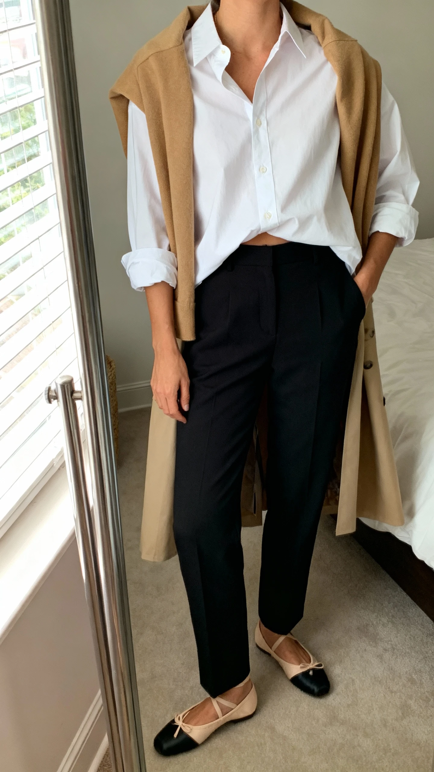 iPhone mirror selfie of a woman wearing an oversized crisp white button-up shirt half-tucked into tailored black trousers, sleeves rolled, with a tan trench draped over shoulders and classic ballet flats, face not visible, cropped at shoulders, bedroom mirror, natural window light, iPhone photo quality.