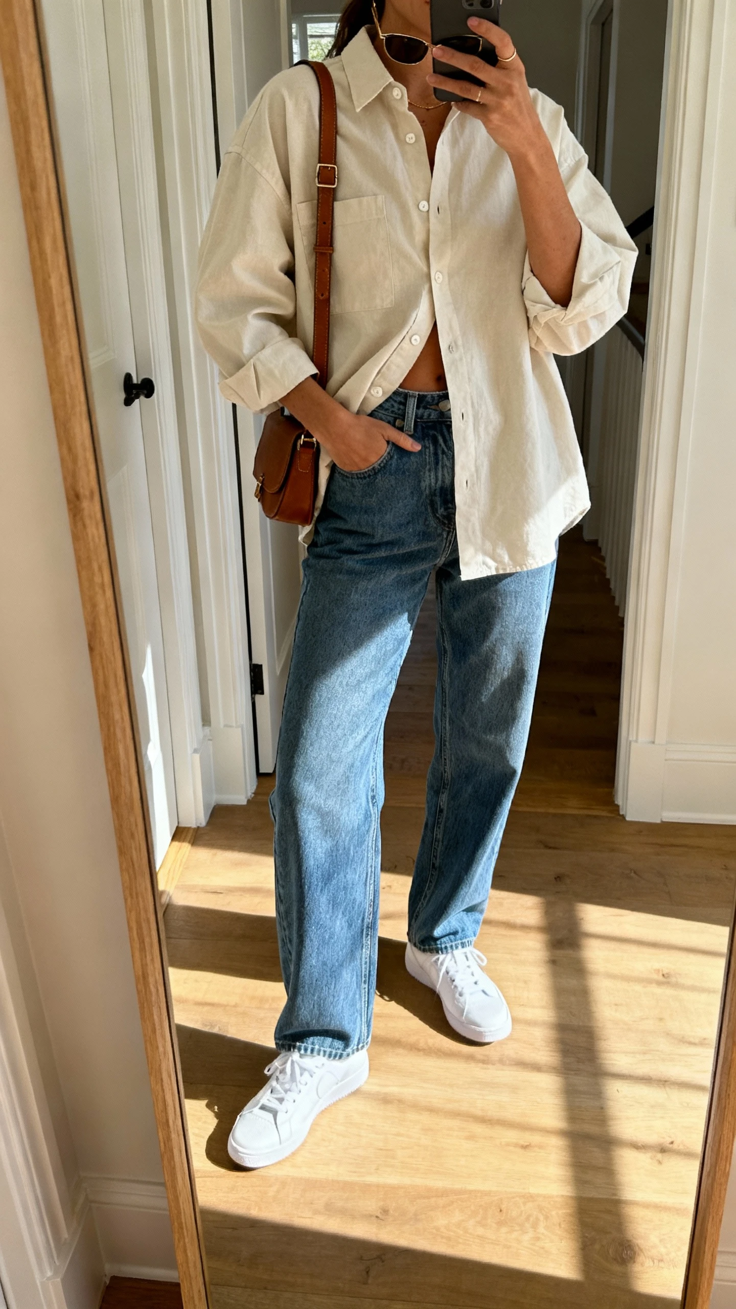 iPhone mirror selfie of a woman wearing an oversized cotton button-down half-tucked into relaxed straight-leg denim, classic white sneakers, a leather crossbody, and simple sunglasses; face not visible, cropped at shoulders, entryway mirror with natural daylight, iPhone photo quality.