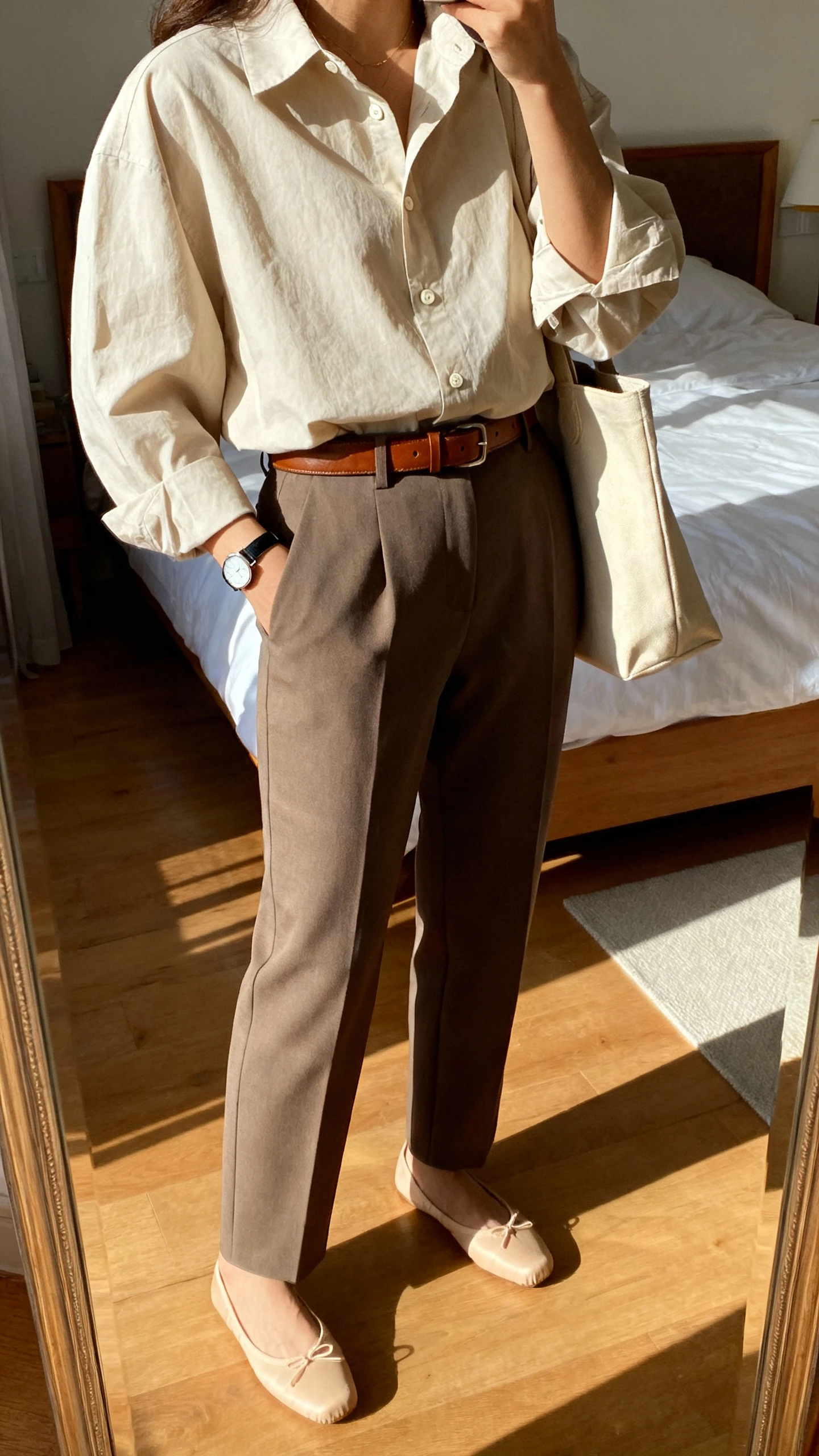 iPhone mirror selfie of a woman wearing an oversized cotton button-down tucked into tailored trousers, leather belt, classic ballet flats, slim watch, tote bag, face not visible, cropped at shoulders, bedroom mirror with warm indoor lighting, iPhone photo quality