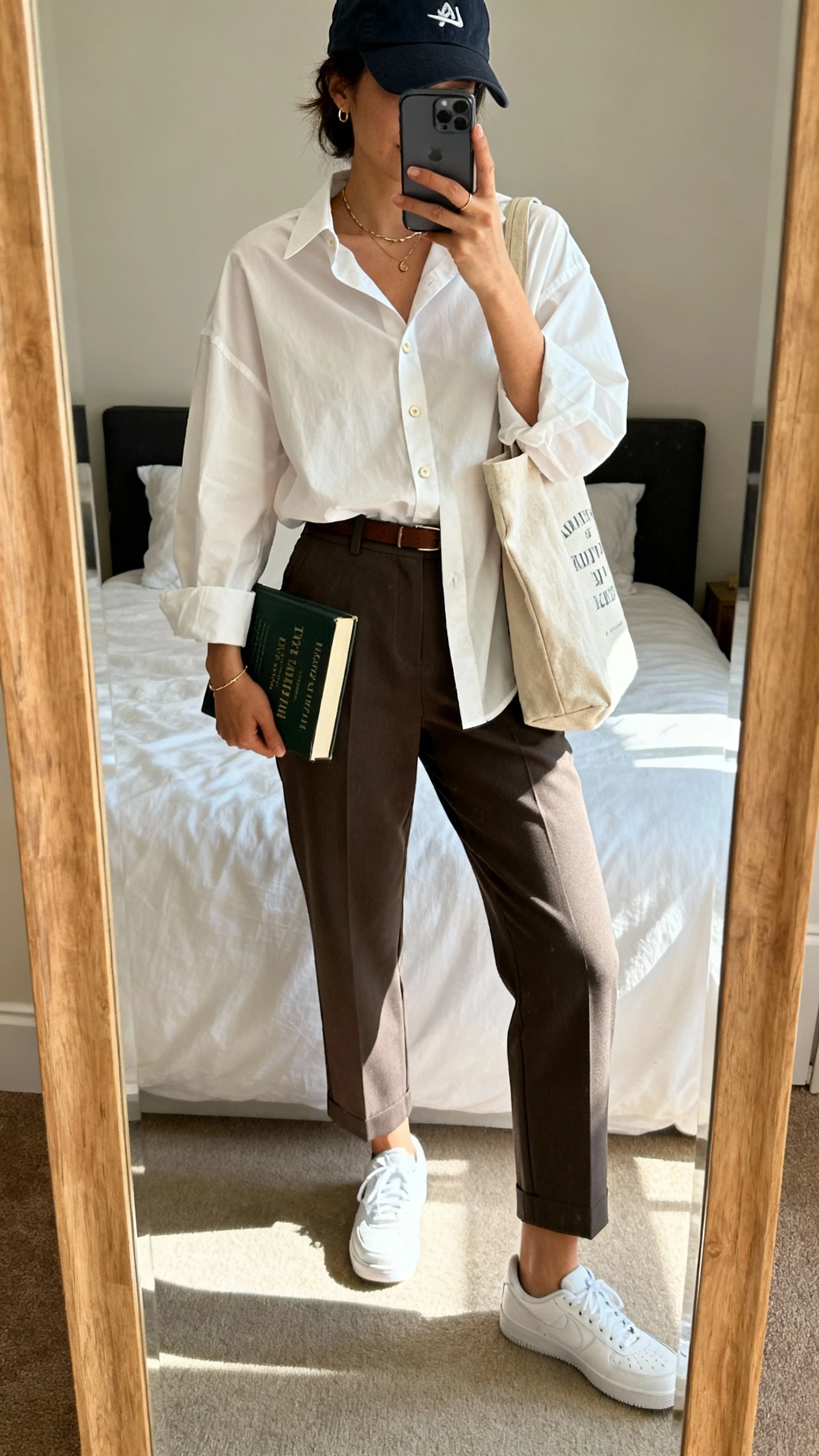 iPhone mirror selfie of a woman wearing an oversized crisp button-down tucked into tailored ankle trousers, white sneakers, baseball cap, and a canvas book tote with minimal jewelry, face not visible, cropped at shoulders, bedroom mirror with natural afternoon light, iPhone photo quality