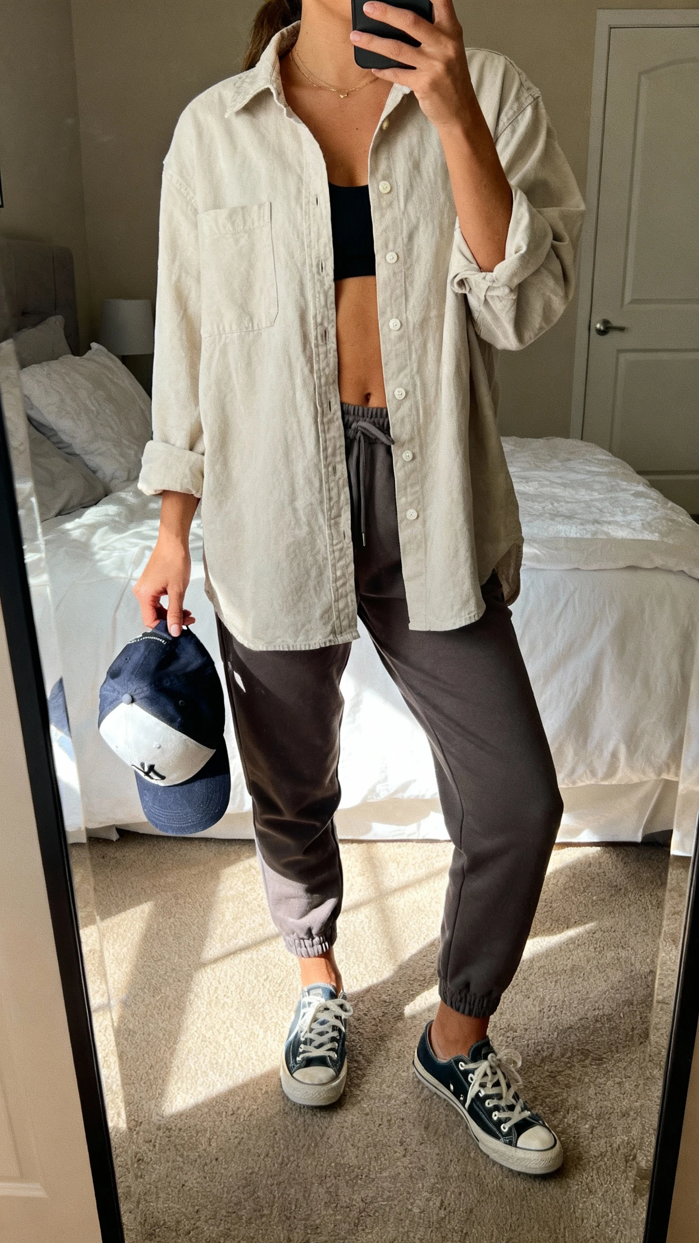 iPhone mirror selfie of a woman wearing an oversized button-up shirt half-tucked into tapered joggers with low-top sneakers and a baseball cap in hand, face not visible, cropped at neck, bedroom mirror, soft afternoon light, iPhone photo quality.