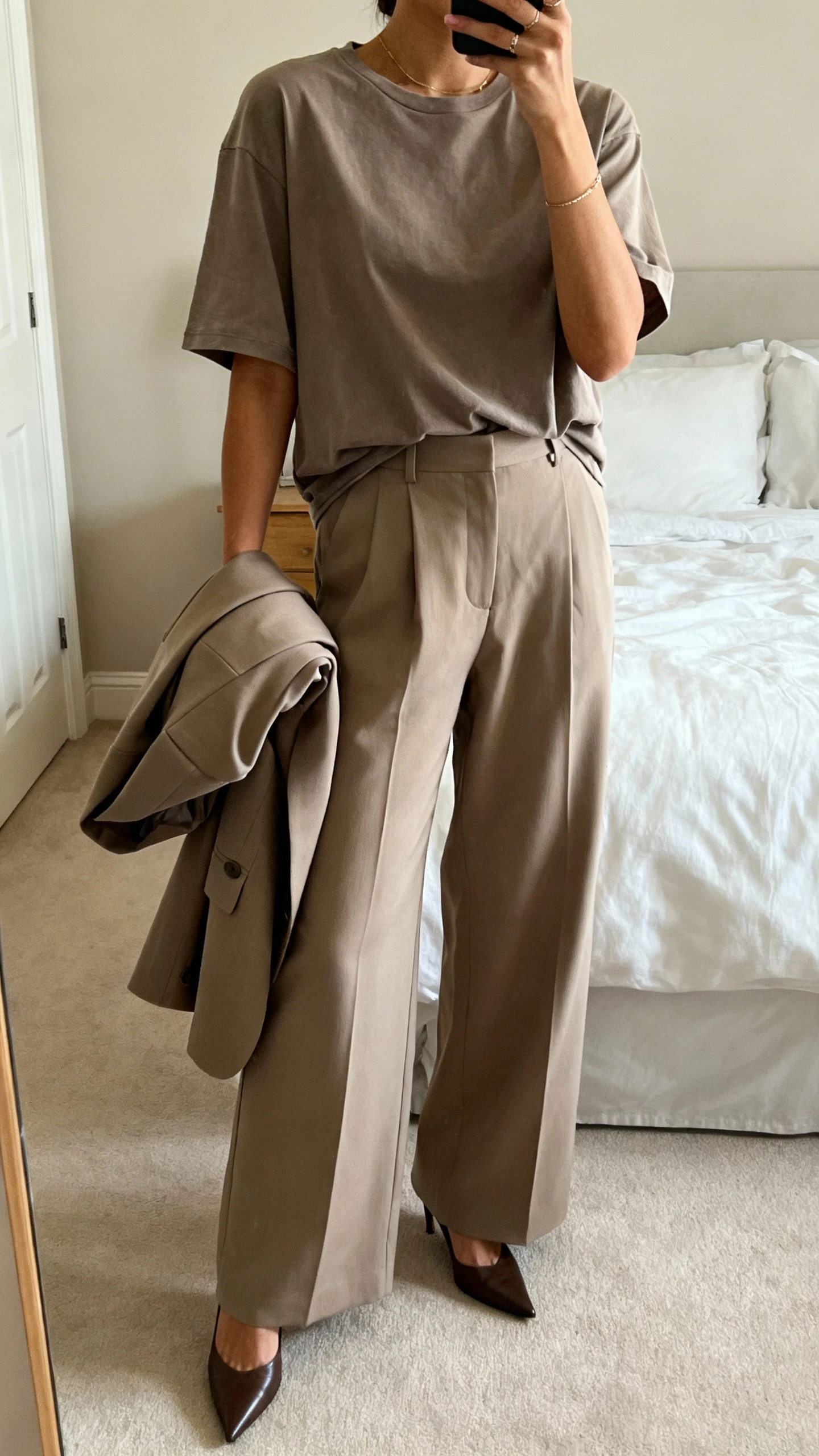 iPhone mirror selfie of a woman wearing an oversized T-shirt with high-waisted wide-leg trousers, pointed-toe heels, a structured blazer draped over the arm, and minimal jewelry, face not visible, cropped at shoulders, bedroom mirror with natural daylight, iPhone photo quality.