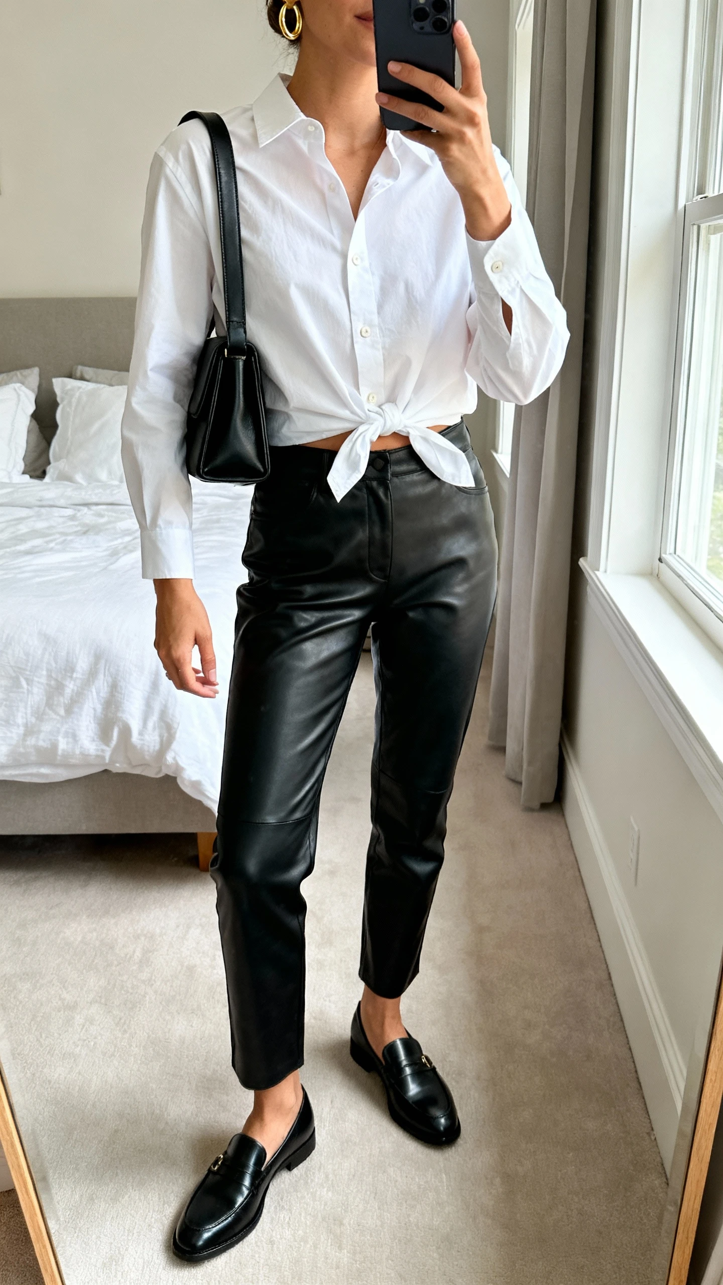 iPhone mirror selfie of a woman wearing black leather pants, a crisp white button-down half-tucked, sleek black leather loafers, simple gold hoops, and a structured black shoulder bag; face not visible, cropped at neck, bedroom mirror, natural window light, iPhone photo quality.