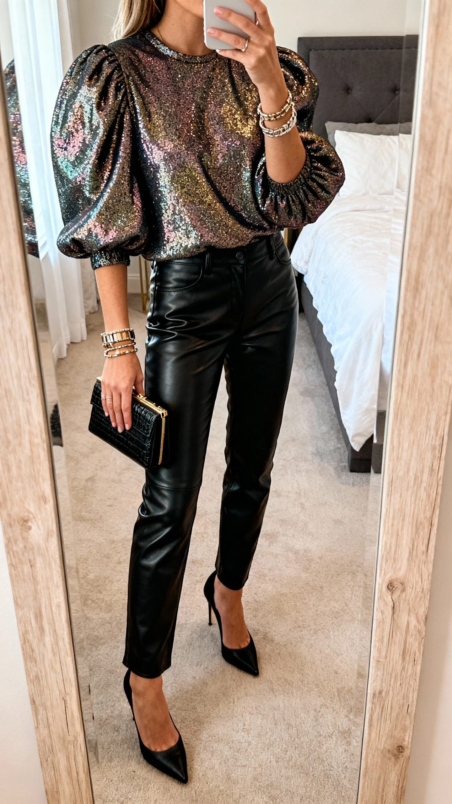 iPhone mirror selfie of a woman wearing black leather pants, a shimmery statement top with puff sleeves, sleek black heels, mini clutch, and layered bracelets, face not visible, cropped at shoulders, bedroom mirror, warm indoor lighting, iPhone photo quality.