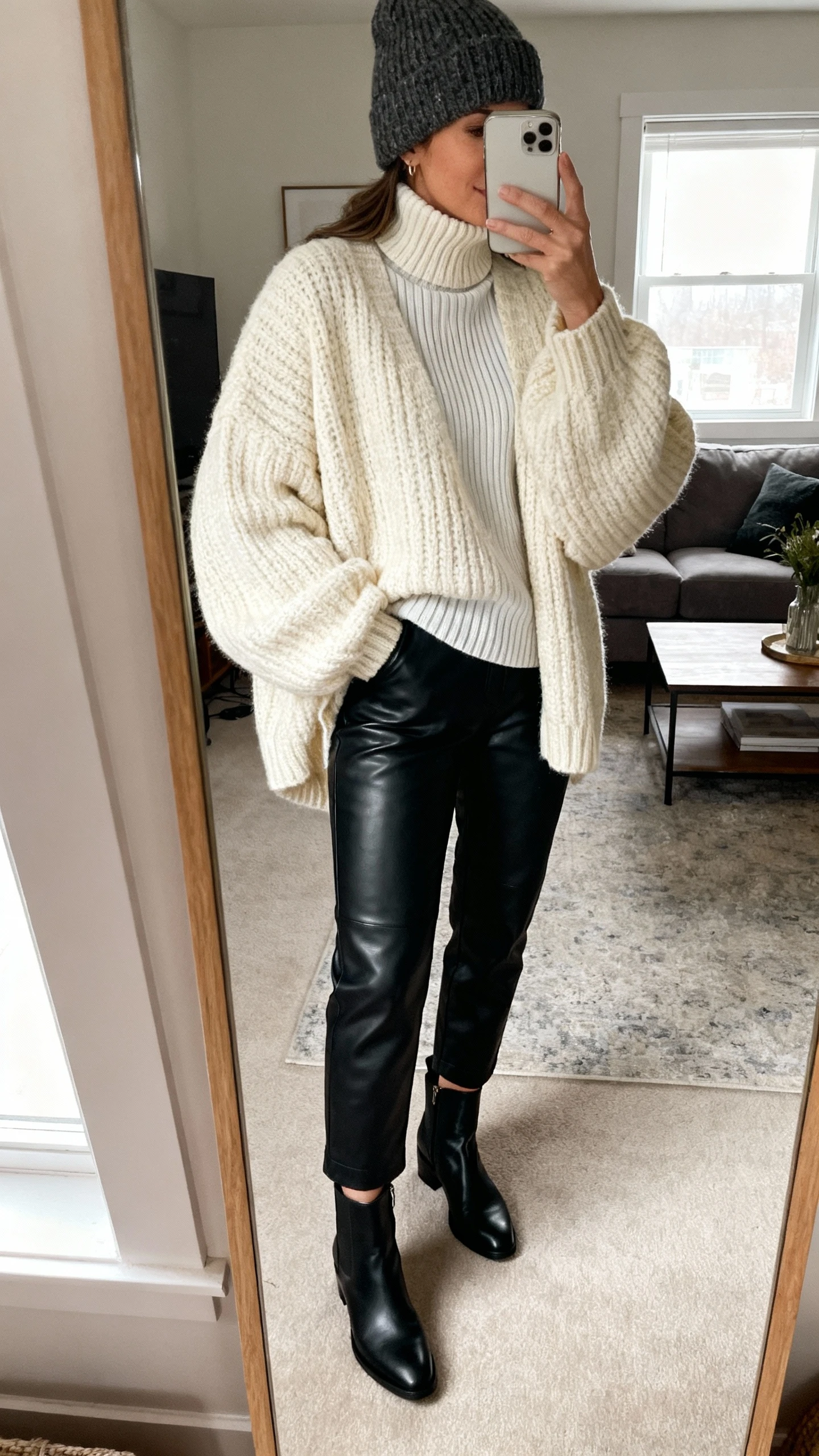 iPhone mirror selfie of a woman wearing black leather pants, an oversized chunky cream sweater layered over a thin ribbed turtleneck, black ankle boots, and a wool beanie; face not visible, cropped at shoulders, living room mirror, soft window light, iPhone photo quality.