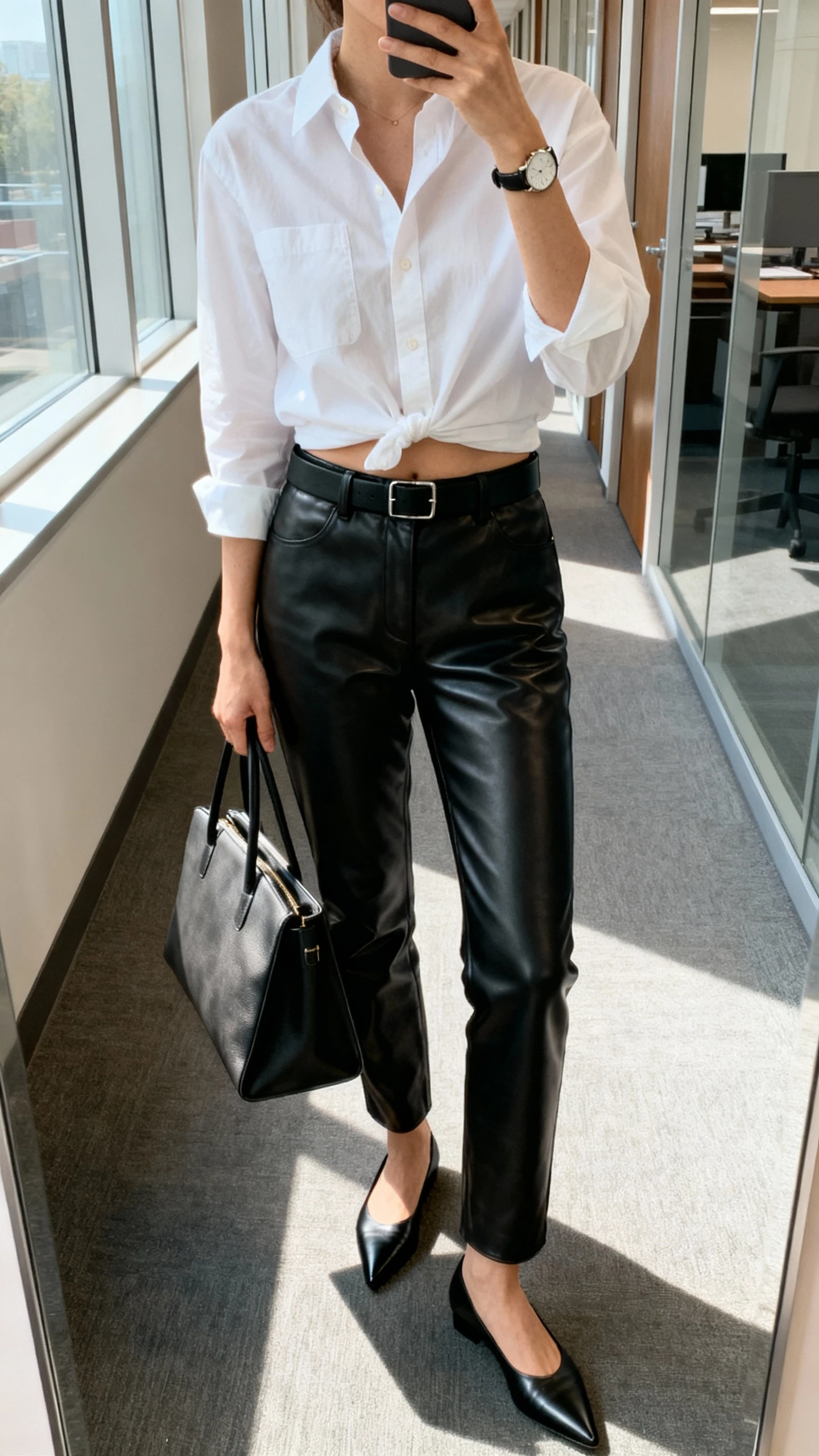 iPhone mirror selfie of a woman wearing black leather pants, a crisp white button-down half-tucked, minimalist black belt, pointed black flats, structured tote, and a simple watch, face not visible, cropped at neck, office hallway mirror, natural window light, iPhone photo quality.