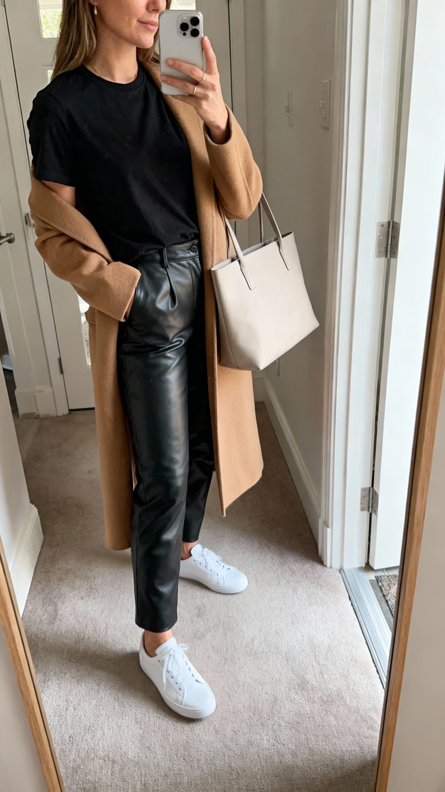 iPhone mirror selfie of a woman wearing black leather pants, a monochrome black tee, clean white low-top sneakers, and a longline camel coat with a minimalist tote; face not visible, side angle, entryway mirror, natural daylight, iPhone photo quality.