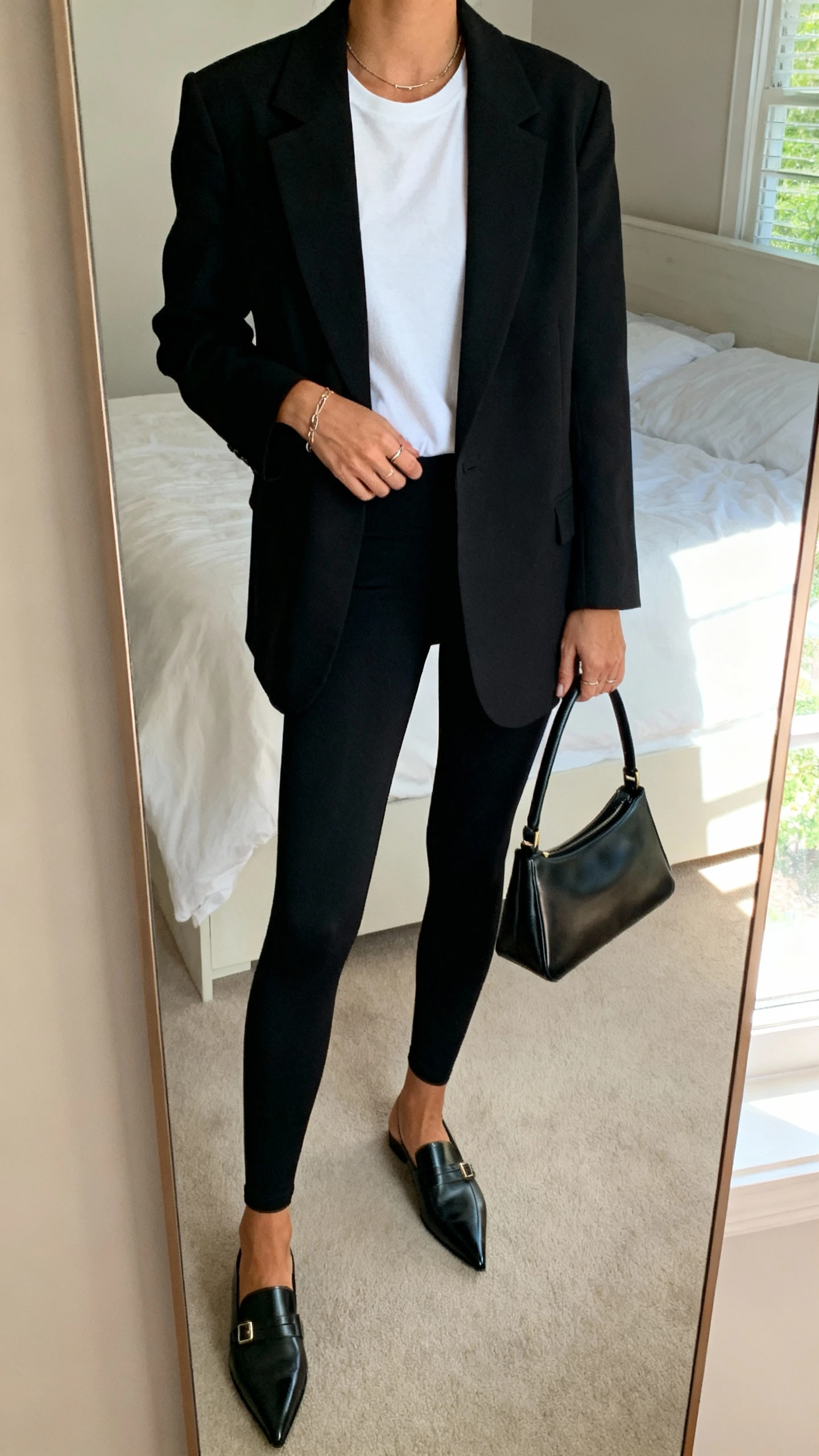 iPhone mirror selfie of a woman wearing black leggings, a structured black blazer over a white tee, pointed loafers, minimalist jewelry, and a sleek top-handle bag, face not visible, cropped at neck, bedroom mirror, natural window light, iPhone photo quality.