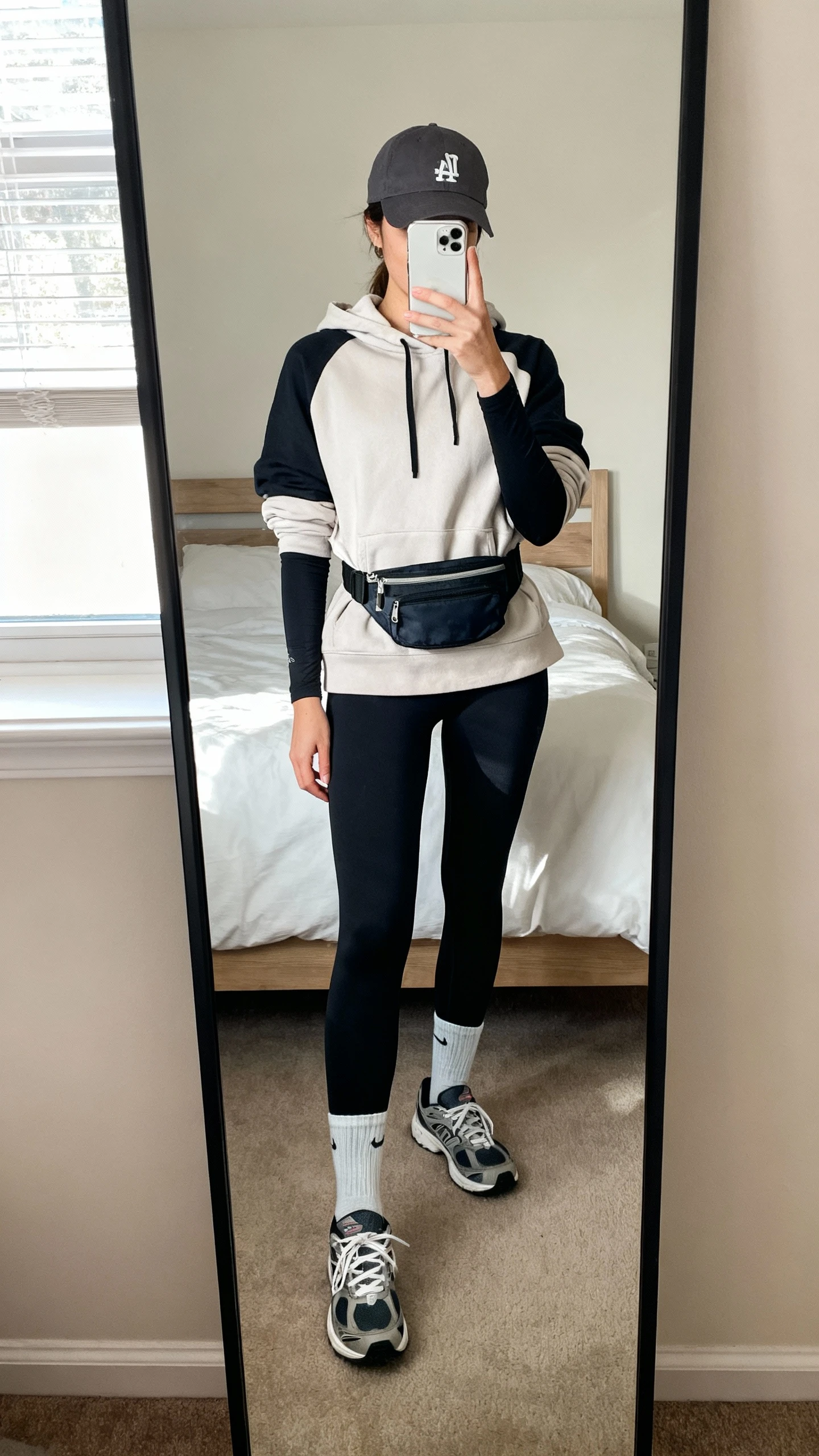 iPhone mirror selfie of a woman wearing black power-leggings, a layered hoodie over a fitted long-sleeve, ankle socks and sporty trainers, plus a crossbody belt bag and baseball cap; face not visible, cropped at neck, bedroom full-length mirror, soft morning window light, iPhone photo quality.