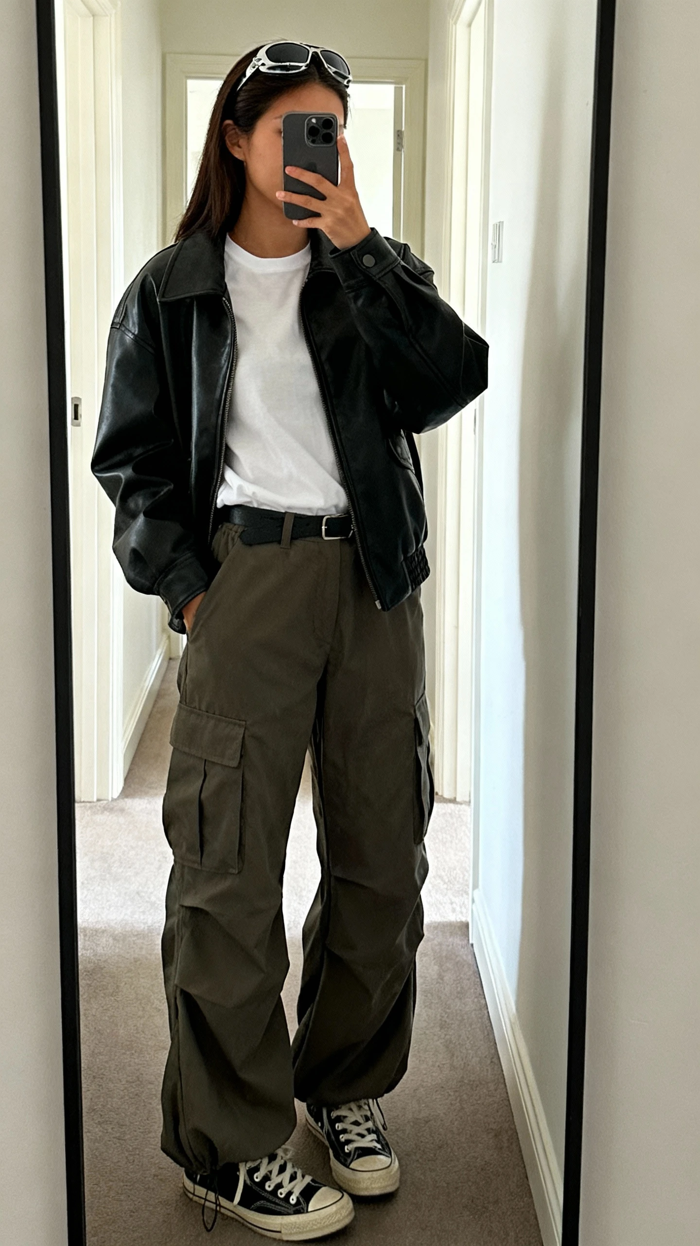 iPhone mirror selfie of a woman wearing cargo parachute pants, a crisp white tee tucked in, and a black leather jacket, with aviator sunglasses perched on head and low-top sneakers, face not visible, cropped at shoulders, hallway full-length mirror, indoor daylight, iPhone photo quality.