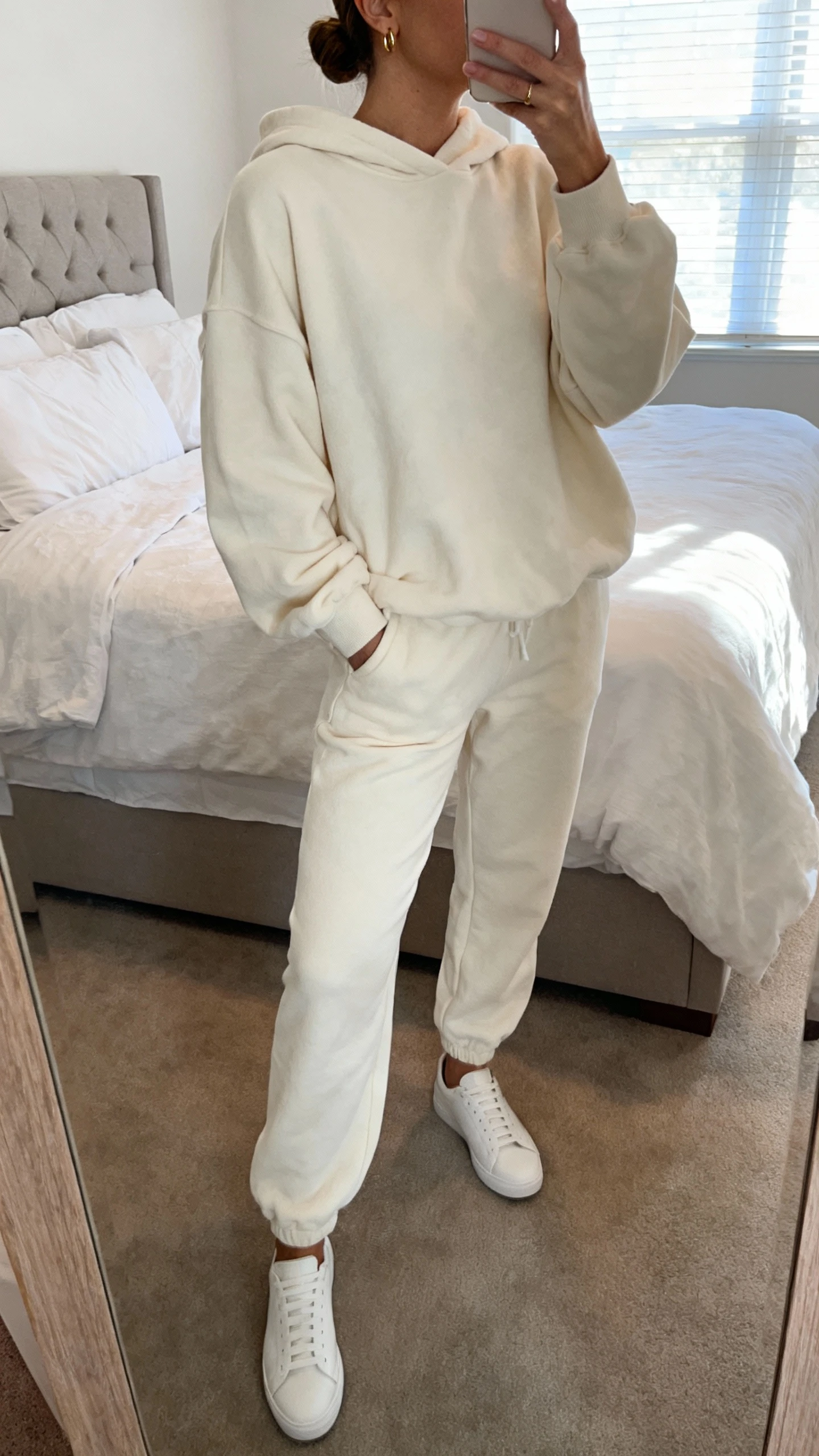 iPhone mirror selfie of a woman wearing cloud-soft matching sweat set (relaxed hoodie and joggers) with sleek white minimalist sneakers, tiny gold hoops, low bun, face not visible, cropped at neck, bedroom mirror with natural morning window light, iPhone photo quality.