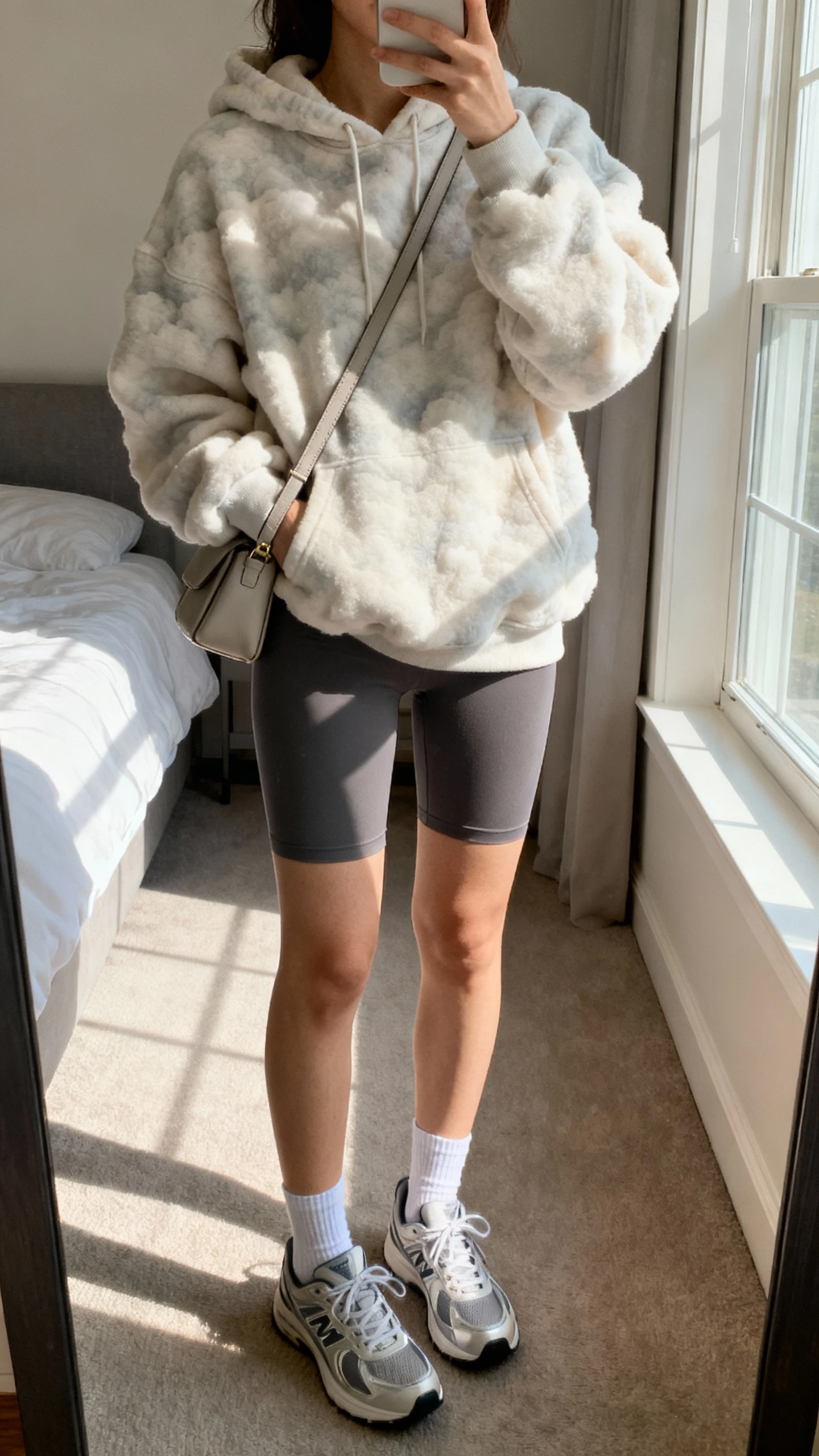 iPhone mirror selfie of a woman wearing cloud-soft hoodie, biker shorts, sleek sneakers, low socks, simple crossbody bag, face not visible, cropped at neck, bedroom mirror with morning window light, iPhone photo quality