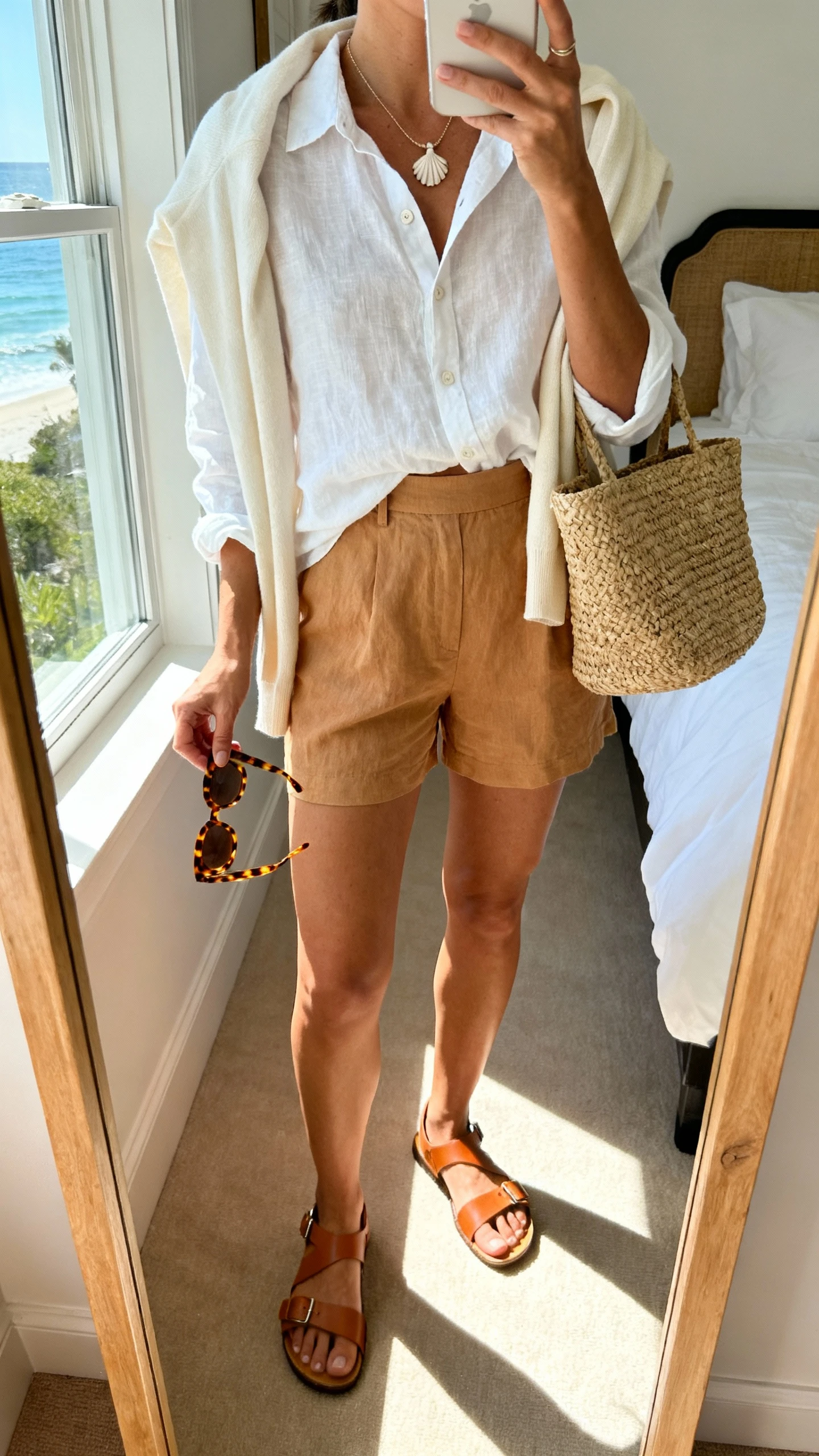 iPhone mirror selfie of a woman wearing coastal cool layers: white linen button-up half-tucked into tan linen shorts, lightweight cream cardigan over shoulders, tan leather sandals, raffia tote, delicate shell necklace, and tortoiseshell sunnies in hand, face not visible, cropped at shoulders, sunlit bedroom mirror near window, iPhone photo quality.