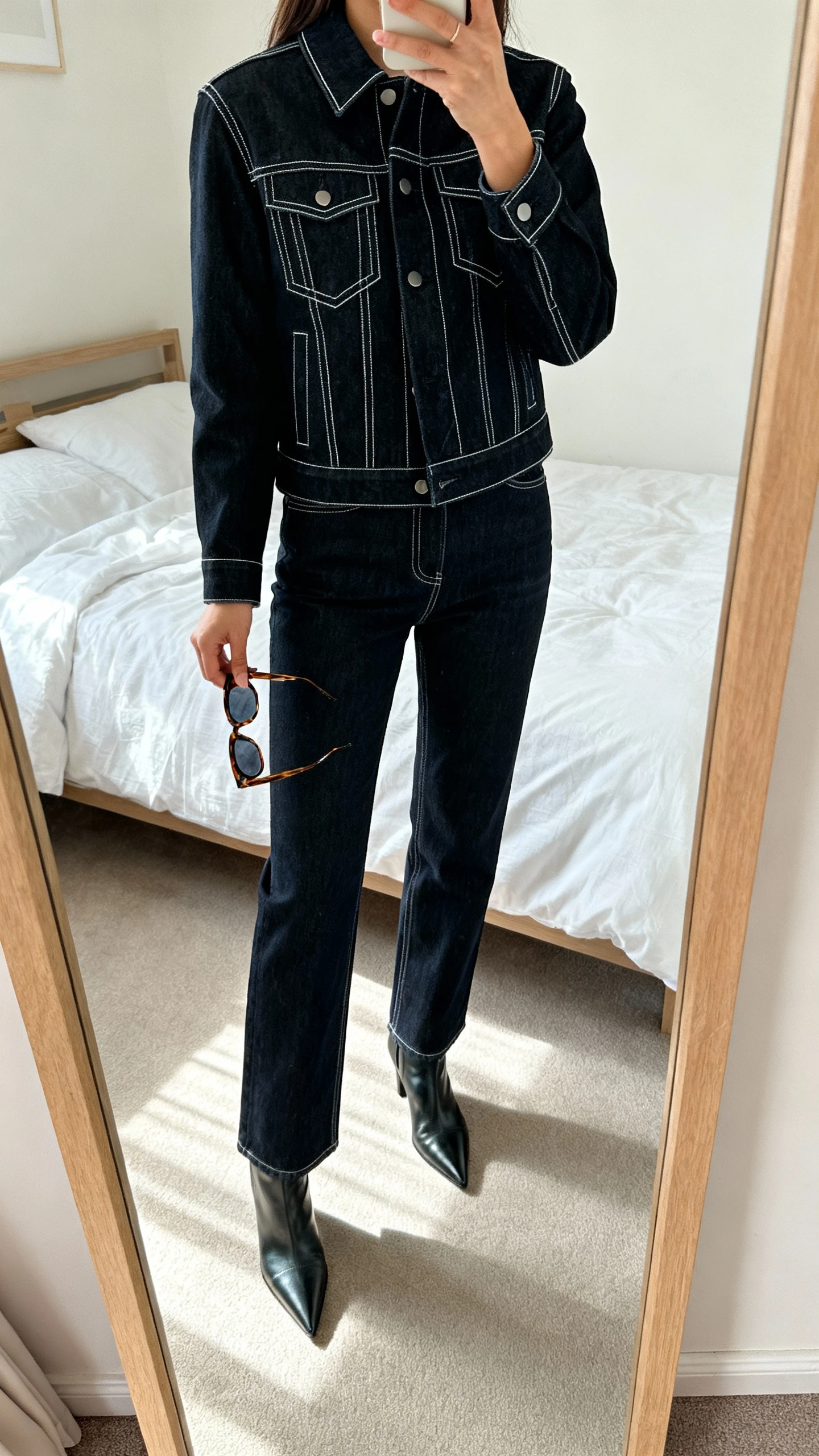 iPhone mirror selfie of a woman wearing denim-on-denim: structured dark-wash jean jacket with sharp lines and matching straight-leg jeans, pointed ankle boots, and angular sunglasses held in hand, face not visible, bedroom mirror with soft daylight, iPhone photo quality.