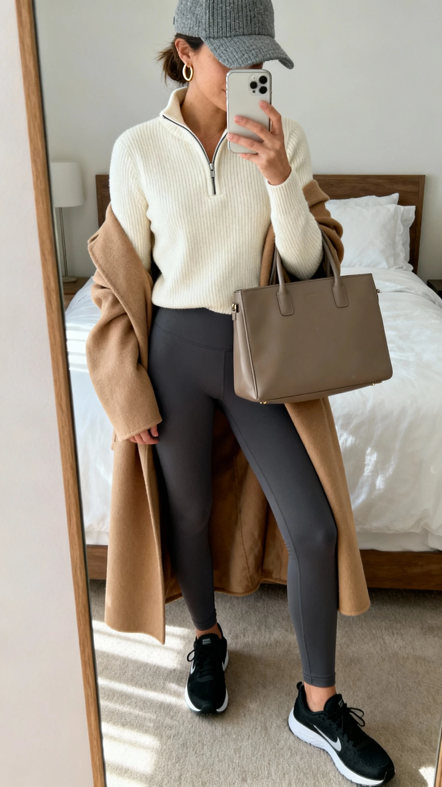 iPhone mirror selfie of a woman wearing elevated athleisure: cream half-zip knit, charcoal high-waisted leggings, sleek black running shoes, gray wool baseball cap, oversized camel coat over shoulders, structured tote, thin gold hoops, face not visible, cropped at shoulders, bedroom mirror, soft afternoon light, iPhone photo quality