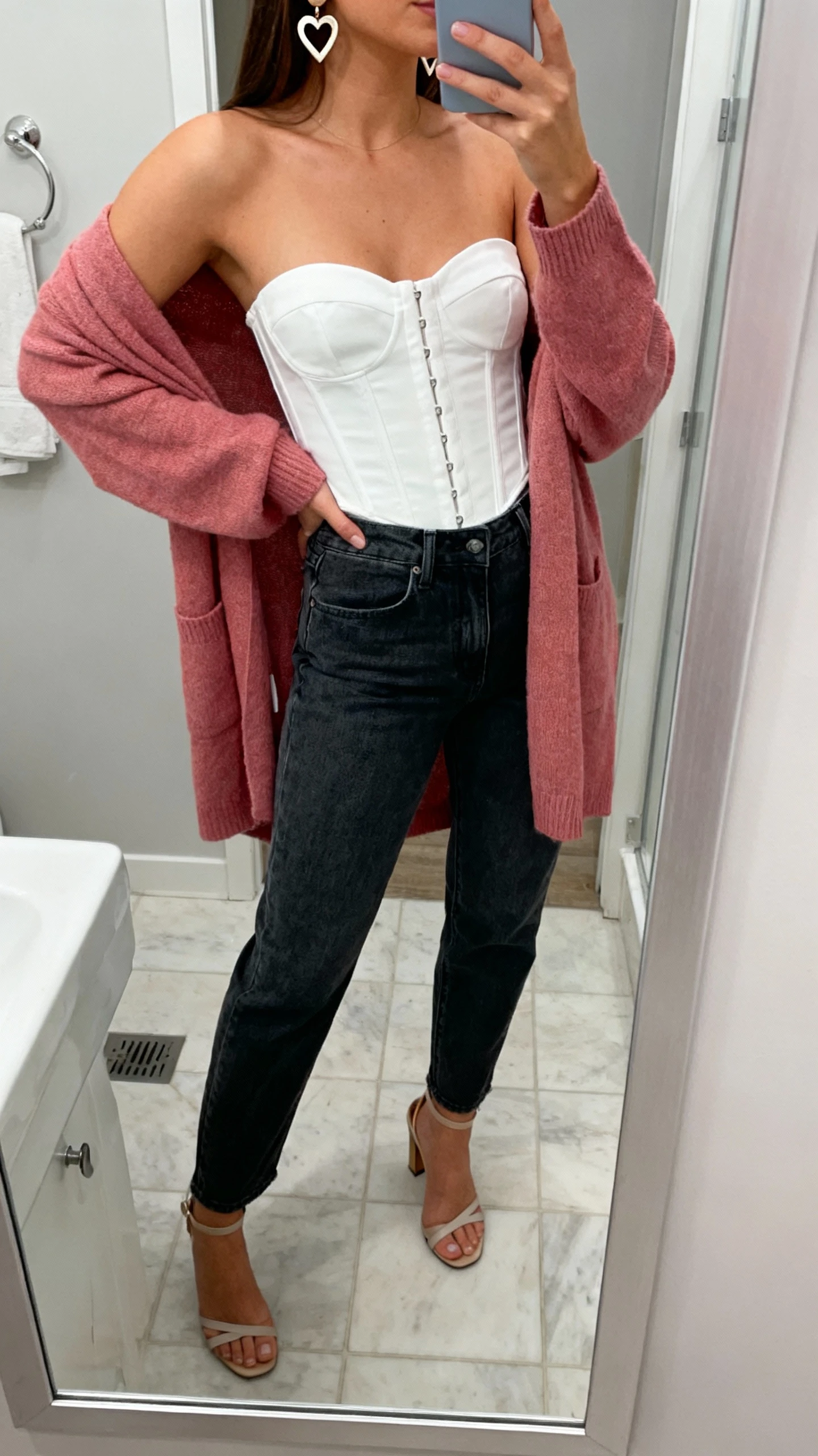 iPhone mirror selfie of a woman wearing elevated dark-wash jeans with a white corset top, rosy pink cardigan draped over shoulders, heart-shaped earrings, and heeled sandals, face not visible, cropped at neck, bathroom mirror with indoor lighting, iPhone photo quality.