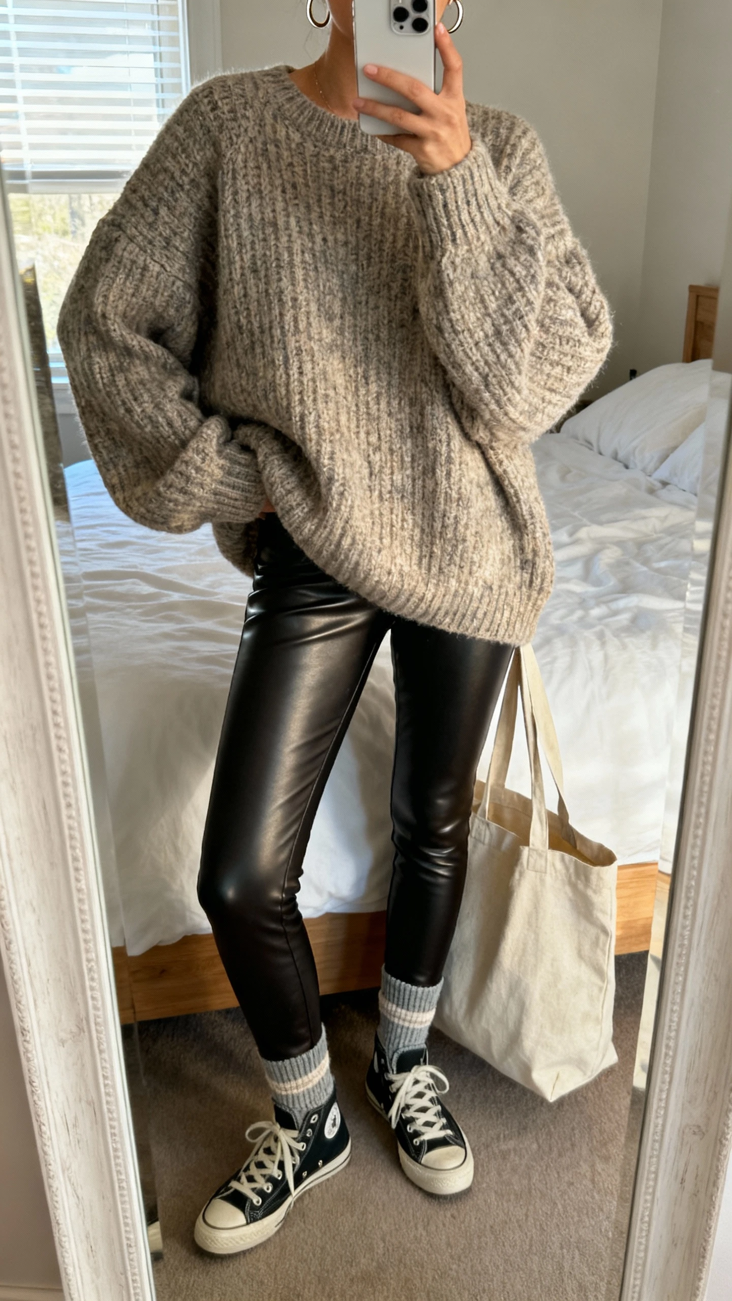 iPhone mirror selfie of a woman wearing faux leather leggings, an oversized chunky knit sweater, crew socks, urban low-top sneakers, a canvas tote, and minimal hoops, face not visible, cropped at neck, bedroom mirror with natural morning window light, iPhone photo quality.