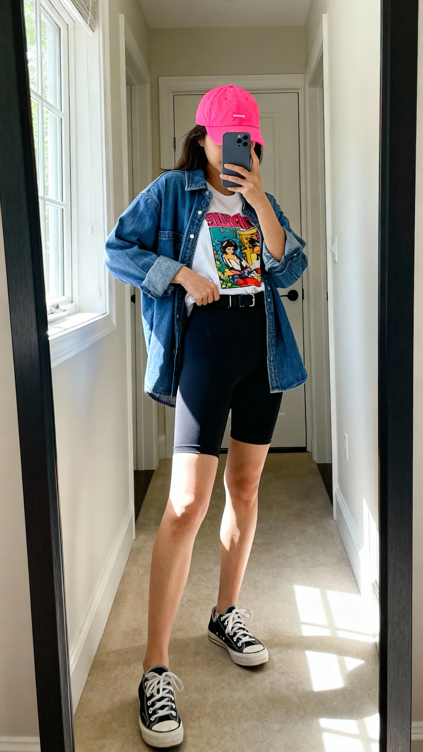 iPhone mirror selfie of a woman wearing graphic tee tucked into biker shorts, denim jacket layered on top, bright color baseball cap for a pop, classic sneakers, face not visible, side angle, entryway mirror with natural window light, iPhone photo quality