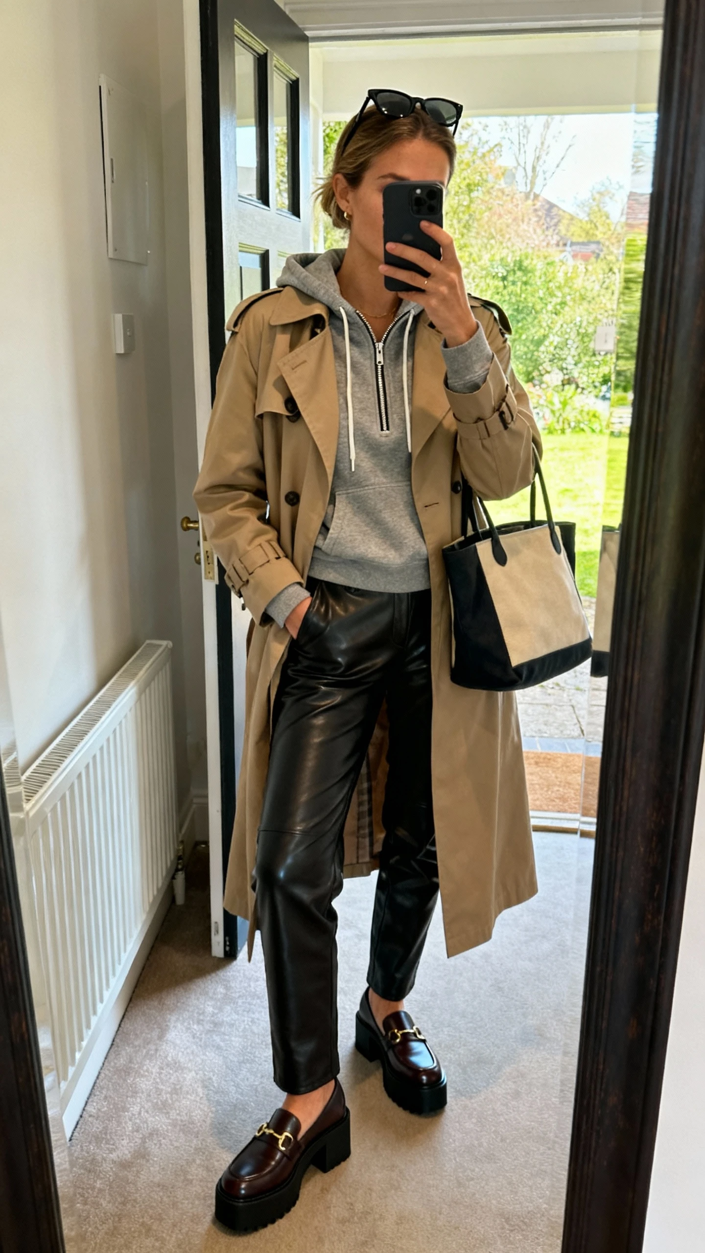 iPhone mirror selfie of a woman wearing leather pants, a zip hoodie layered under a trench coat, and platform loafers, tote bag and sunglasses pushed up on head, face not visible, cropped at shoulders, entryway mirror with daylight, iPhone photo quality.